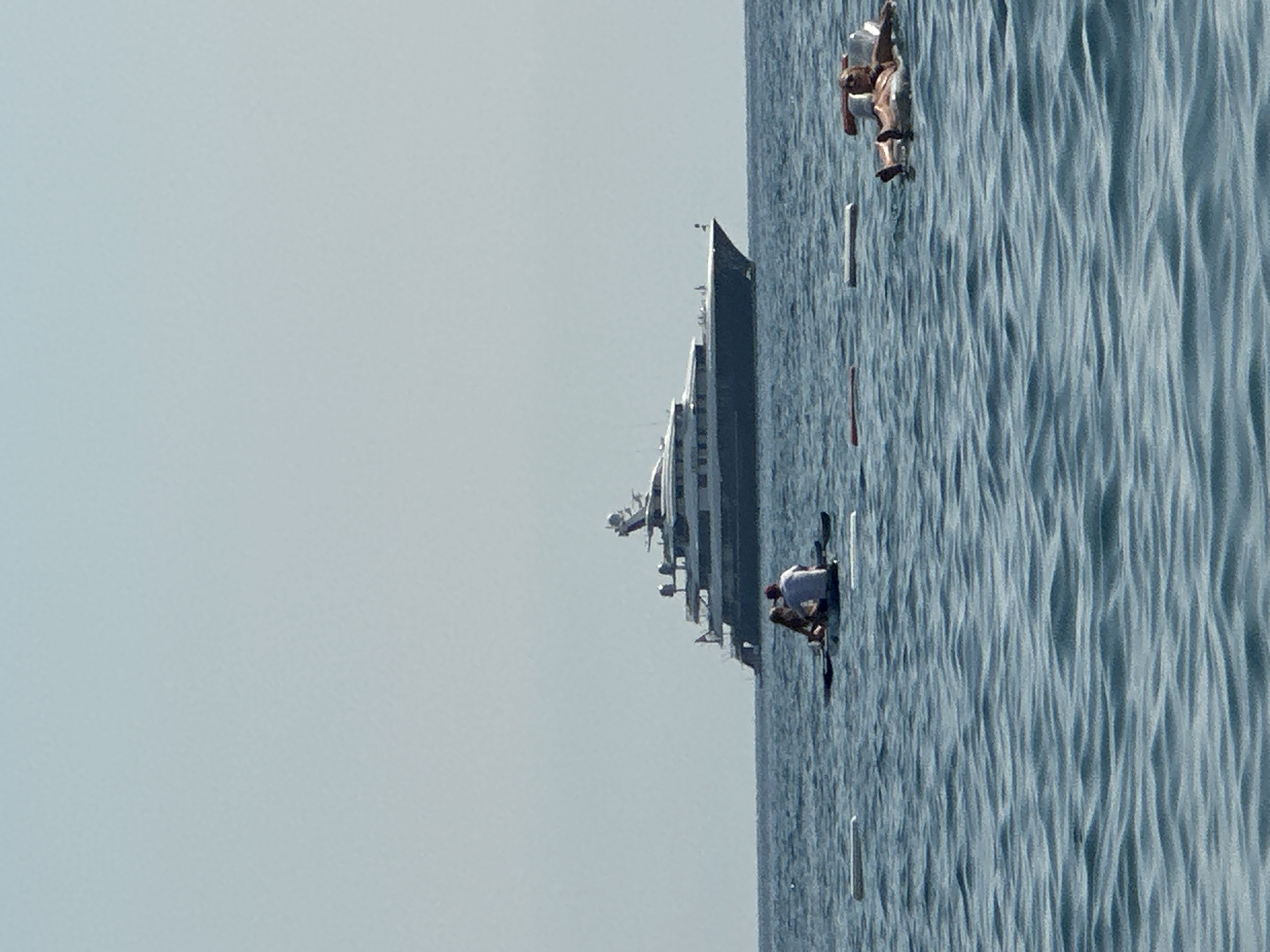 In the waters off the coast of Boreti, Montenegro, a large, multi-decked yacht with a dark hull and white superstructure is anchored in the distance. Closer to shore, two people are seated on a paddleboard. To the right, another individual reclines on a white inflatable raft. Several other white floating objects are scattered across the water's surface. The sea is a deep blue with gentle waves, and the sky is a hazy light blue, suggesting a clear, sunny day. The overall scene depicts a leisurely day at sea, with people enjoying various forms of water recreation.