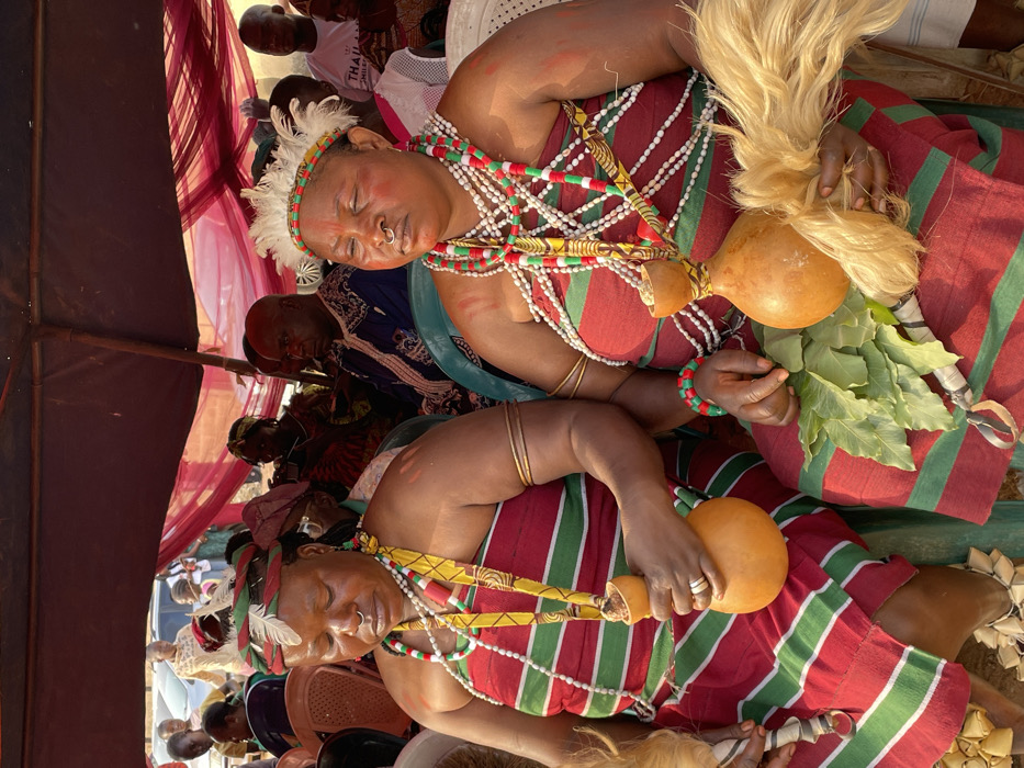 The image captures a vibrant cultural gathering, likely an outdoor ceremony, under what appears to be a red canopy or tent. Two women are prominently featured in the foreground, seated, their eyes closed in what could be a moment of rest, meditation, or spiritual trance.
Both women are adorned in elaborate traditional attire. They wear matching red and green striped wrap skirts or dresses. The woman in the upper part of the frame also wears a solid red top, while the woman in the lower part of the frame has her upper body covered by her multiple necklaces. Their skin tone is rich and dark, and both have what appear to be red ritualistic markings on their faces and shoulders. Each woman wears a nose ring and a headband decorated with white feathers and red and green beads.
Numerous intricate necklaces drape around their necks, fashioned from strings of red, white, and green beads. Some of these necklaces are interspersed with yellow patterned fabric or ribbon. They also wear multiple golden bangles on their arms and bracelets made of matching red and green beads.
Distinctive gourds are central to their appearance and possible activity. The upper woman holds a large, light brown gourd adorned with a voluminous tuft of blonde or light-colored fibrous material, possibly hair or straw. Next to her right hand, a cluster of large green leaves is visible. The lower woman holds a smaller, similar gourd in her left hand, which is also decorated with fabric or fiber. Another gourd lies near her feet. She also wears rings on her fingers.
In the background, several other individuals, mostly men, are visible, seated or standing, observing the scene. They are largely out of focus, but some wear traditional clothing. One person in the mid-background wears a white t-shirt with partially visible text that appears to read "THAILAND". The lighting is bright and natural, indicating it is daytime. The overall atmosphere suggests a significant cultural or spiritual event, rich with tradition and community presence.