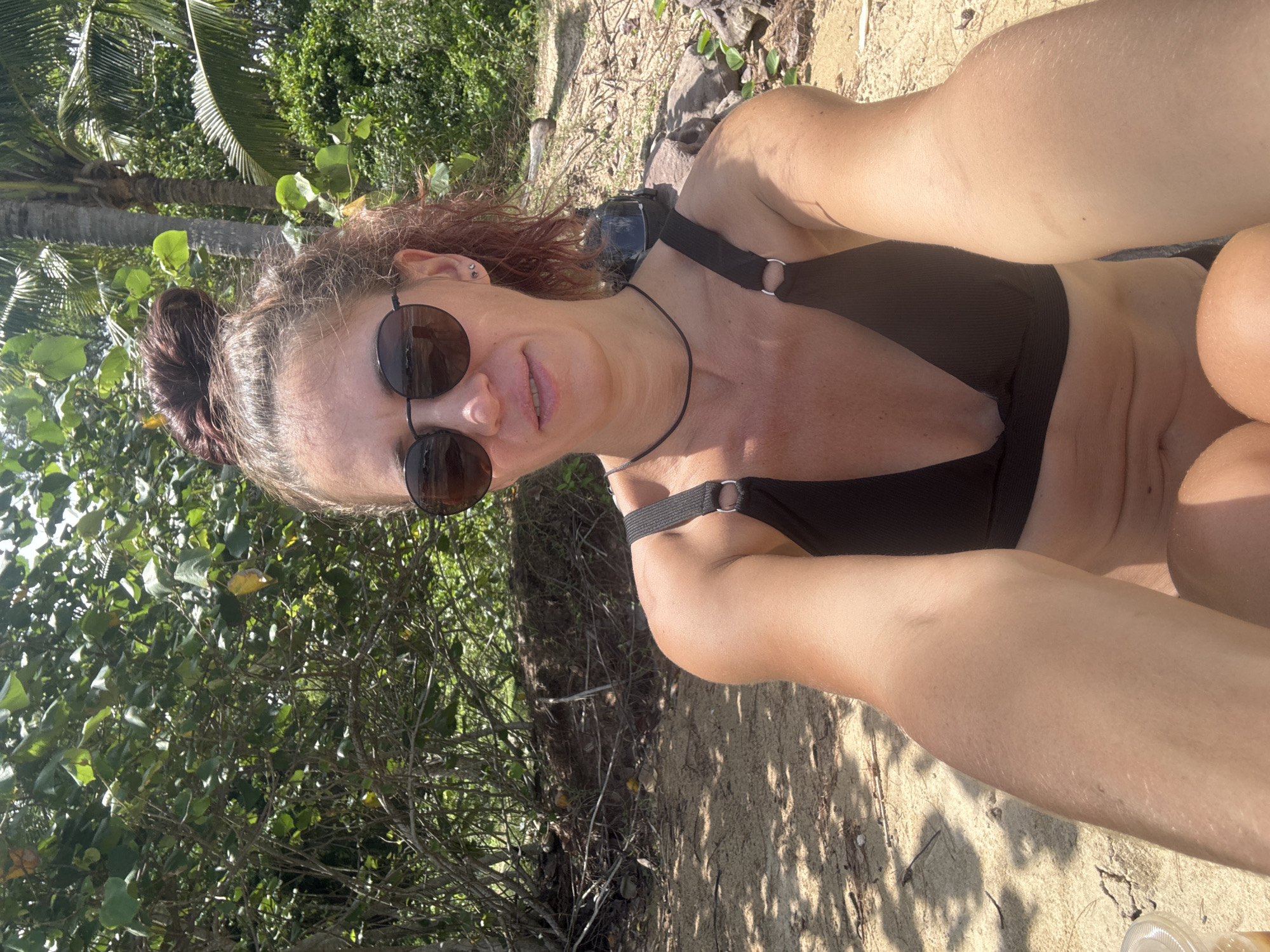 A woman is taking a selfie on a beach. She is wearing a black bikini and sunglasses. Her hair is pulled up in a bun, and she has a slight smile on her face. The background shows lush green foliage, palm trees, and sandy ground with some scattered rocks and debris. The lighting suggests it is a sunny day. The image is taken from a high angle, looking down at the woman.