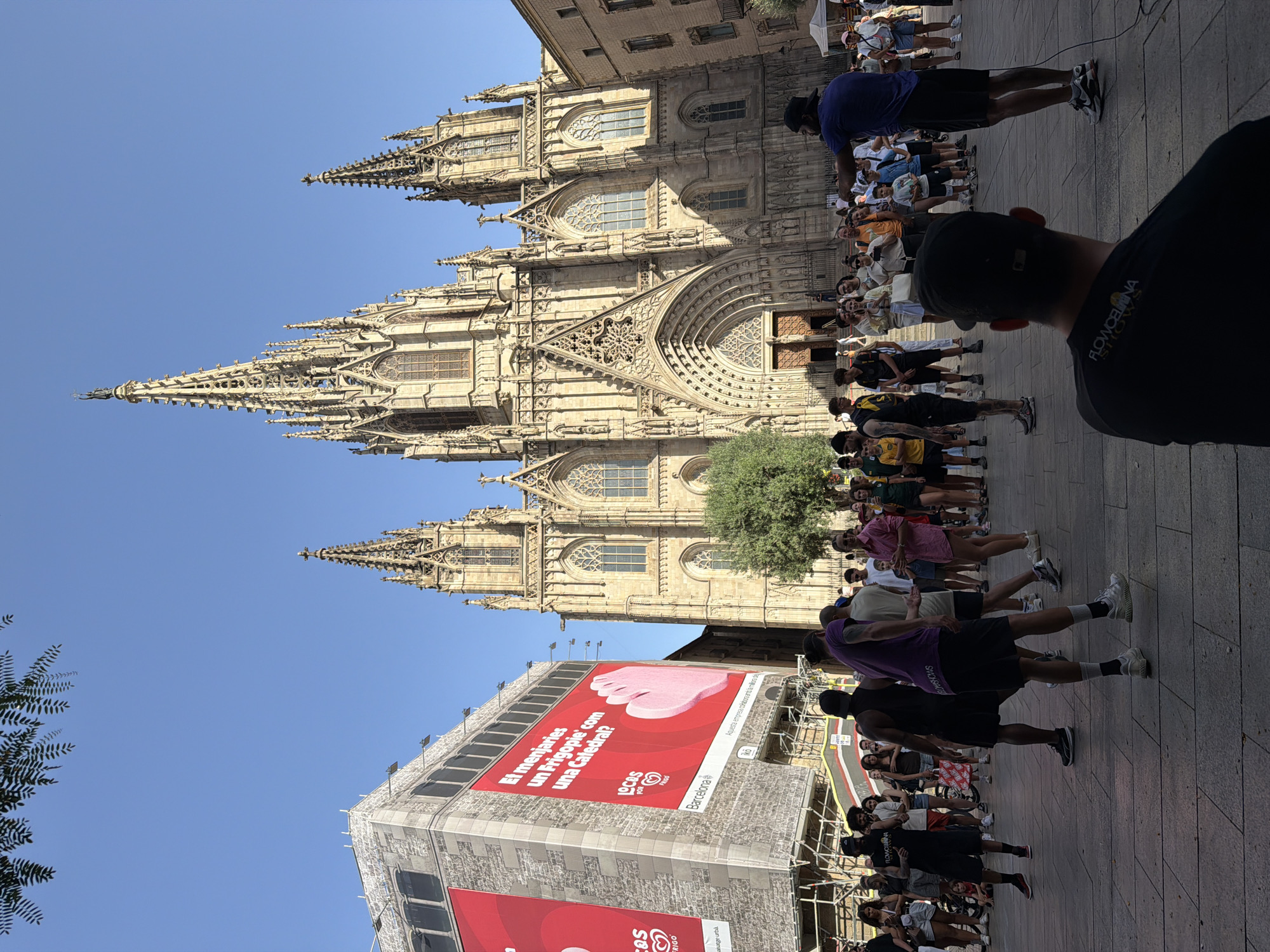 This is a vertical, eye-level, medium shot of a street scene in Barcelona, Spain. The Barcelona Cathedral dominates the background, its gothic spires and intricate stonework silhouetted against a clear blue sky. In the foreground, a crowd of people is gathered on a cobblestone plaza. Some are standing in neat rows, appearing to participate in or observe an organized activity, possibly a dance or performance, led by a man in a purple shirt. Others are standing more casually, watching the proceedings or enjoying the day.
A large red banner is prominently displayed on a building to the left, featuring an image of a pink ice cream and text in Catalan that translates to "Would you eat a Frigopie like a Cathedral?". This suggests a promotional event or advertisement related to ice cream, likely a Frigopie brand, with a playful comparison to the iconic cathedral. The "LOCOS POR FRIGO" (Crazy for Frigo) and "Barcelona" text further confirm the context.
The time of day appears to be daytime, with bright sunlight casting shadows and illuminating the scene, indicating fair weather. The overall atmosphere seems lively and engaging, with a mix of locals and possibly tourists enjoying the urban spectacle and the architectural grandeur of the cathedral. The presence of people dressed in casual summer attire suggests a warm day.