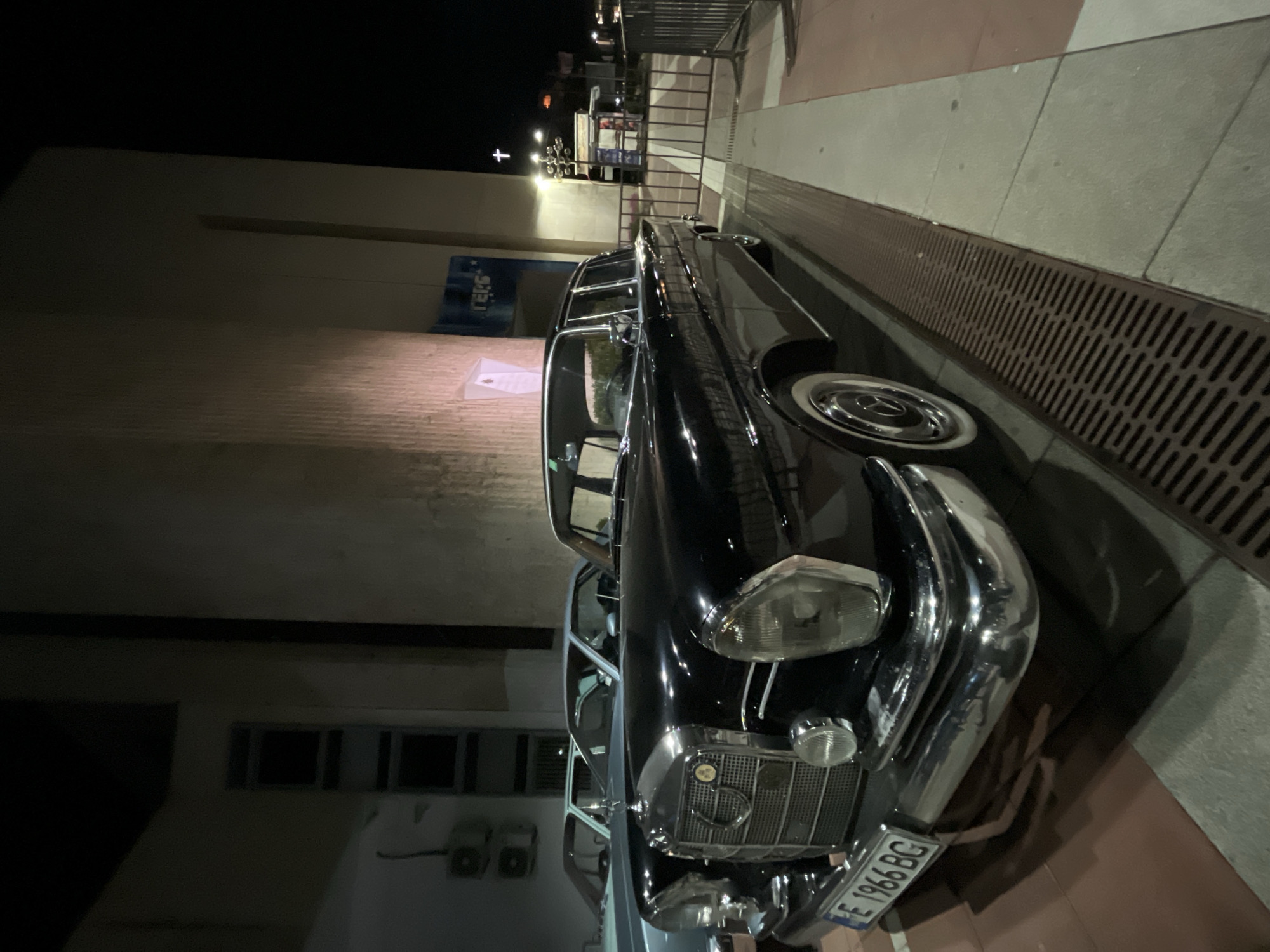 A vintage black Mercedes-Benz sedan is parked on the side of a street in Blagoevgrad, Bulgaria. The car's license plate reads "E 1966 BG". The street is paved and has a metal grate along the curb. A building is in the background, and a metal fence and a cross are visible further down the street. The scene appears to be at night or in low light.