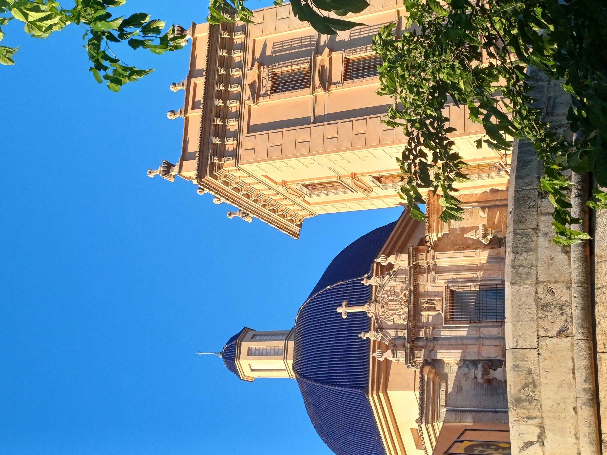 The image shows the Church of Santos Juanes in València, Spain, viewed from a low angle, emphasizing the architectural details against a clear blue sky. Part of the church's blue-tiled dome and bell tower are visible. Green foliage is present in the upper corners of the frame, partially obscuring the view. The building exhibits a mix of architectural styles, with detailed carvings and decorative elements on its facade. Shadows and light play across the textured surfaces of the building.