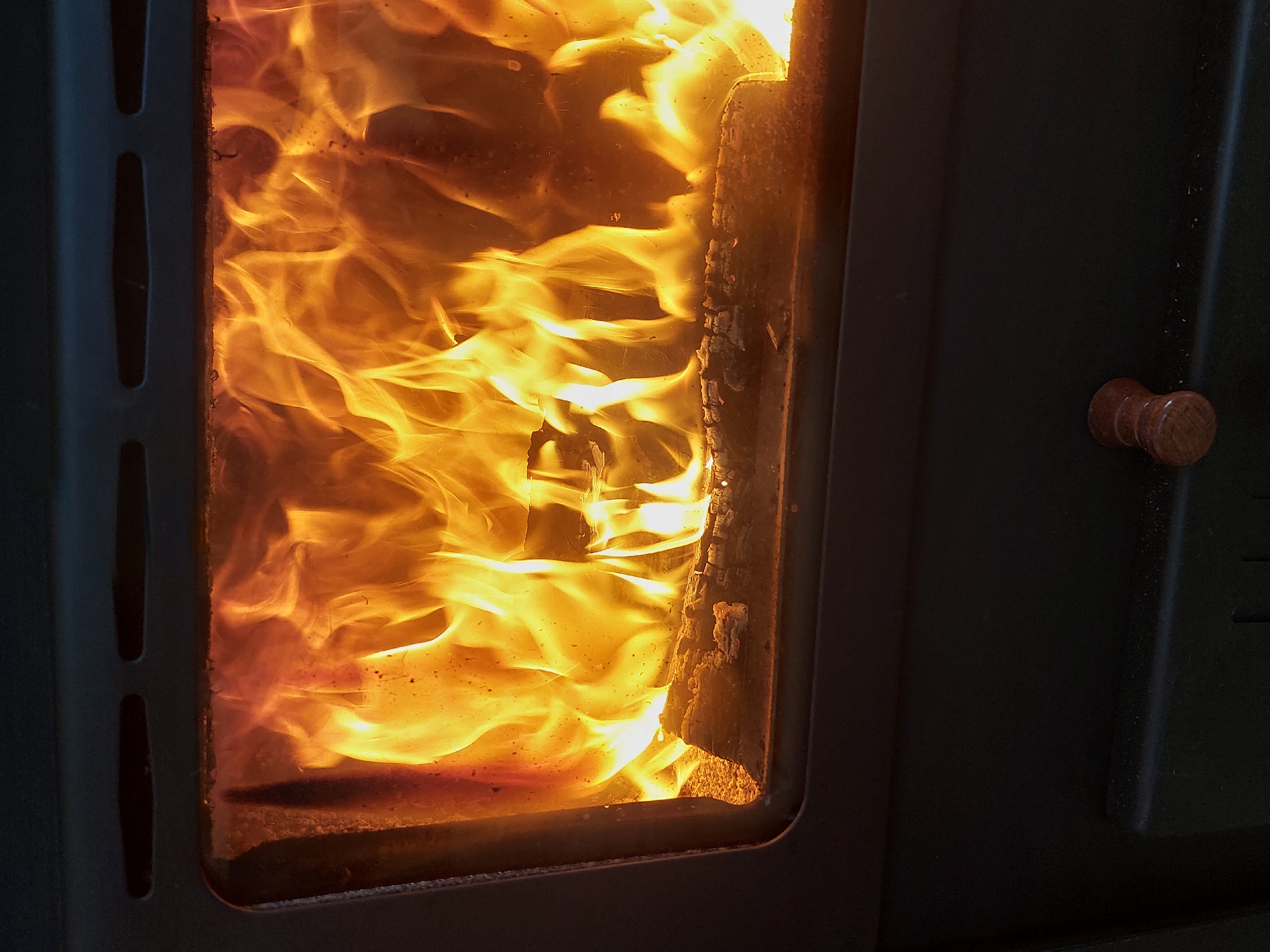 The image shows a wood-burning stove with flames visible through the glass door. A single log is burning. The stove is black, with a wooden handle on the door. The location is not discernible from the image.
