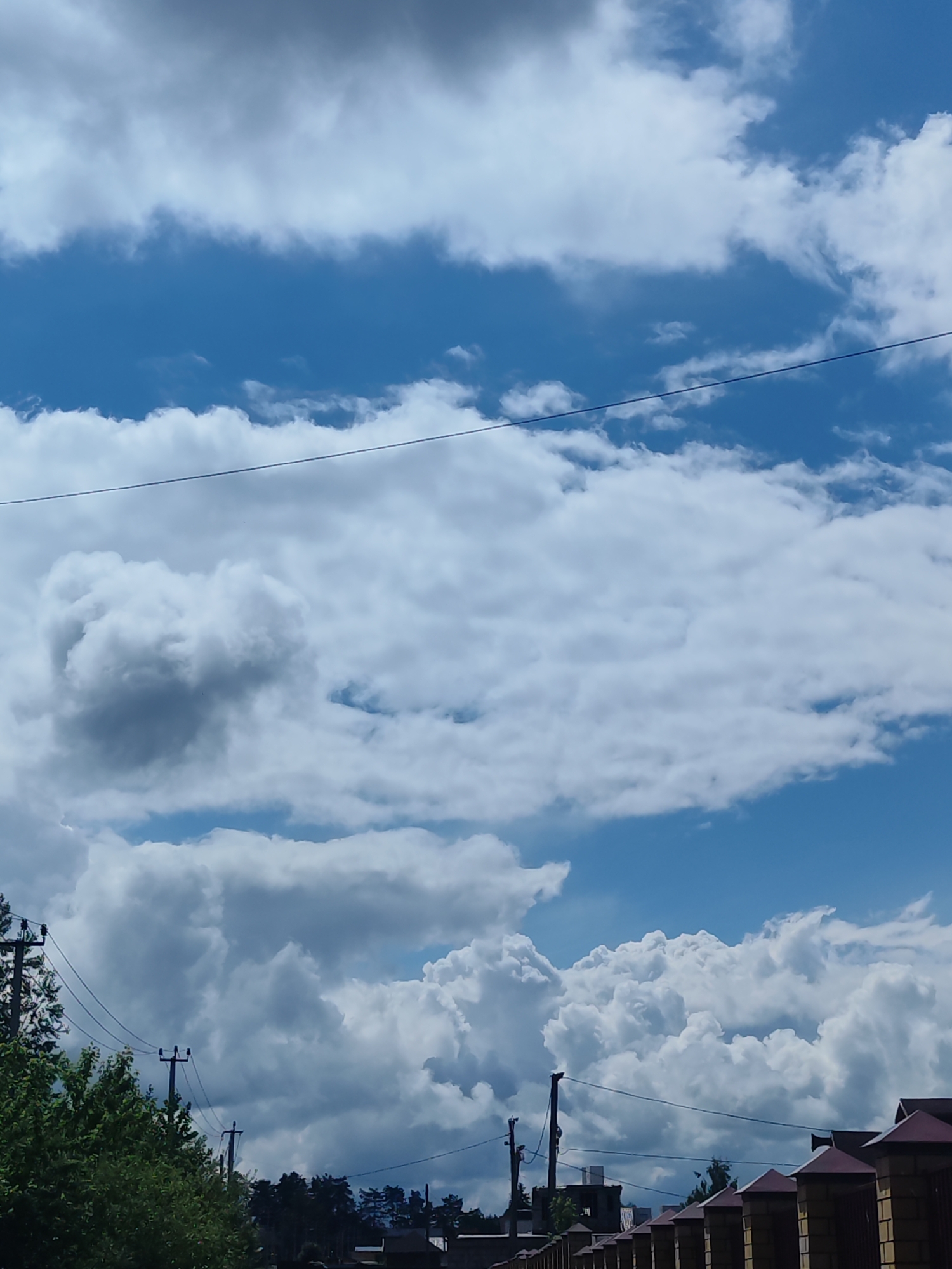 The image presents a bright, daytime scene under a cloudy, blue sky. Several layers of cumulus clouds dominate the upper portion of the view, with visible clear blue sky between and above them. An electrical wire stretches horizontally across the upper section of the image.
In the lower portion, a mix of natural and built elements is visible. Trees and foliage occupy the left side, with telephone poles and wires extending upwards. Further back, silhouettes of residential buildings and denser tree lines are discernible. On the right, a brick fence with dark-red pyramid-shaped caps runs along the edge of a property. The location appears to be a residential area, potentially suburban, within the city of Unknown, Unknown.