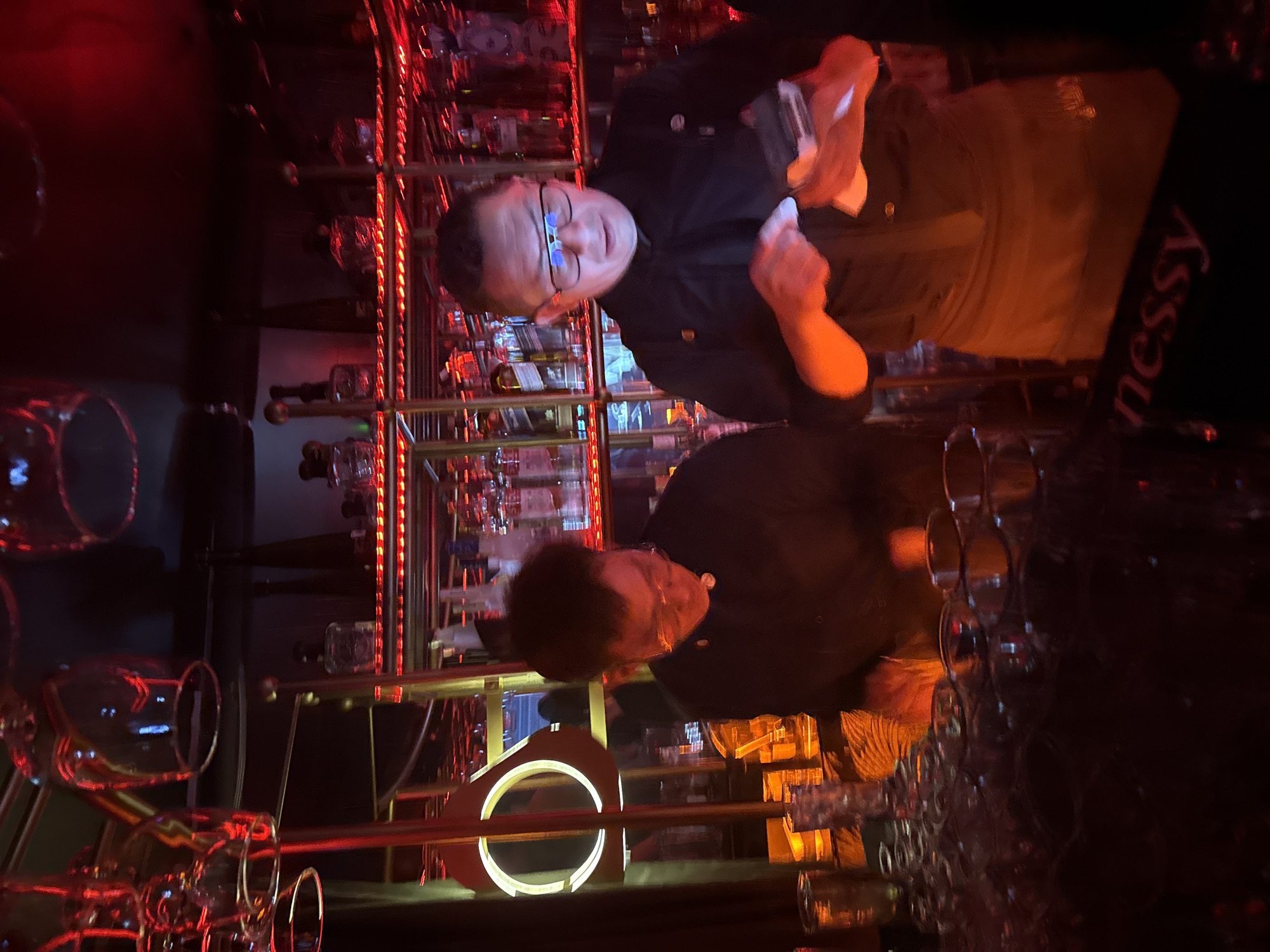 The image shows a bartender preparing a drink at a bar in Mexico City, Mexico. The man wears glasses and a black shirt. A partially visible apron shows the word "nessy." A bar with numerous liquor bottles behind the bartender is lit up with red lights. Wine glasses are in the foreground.