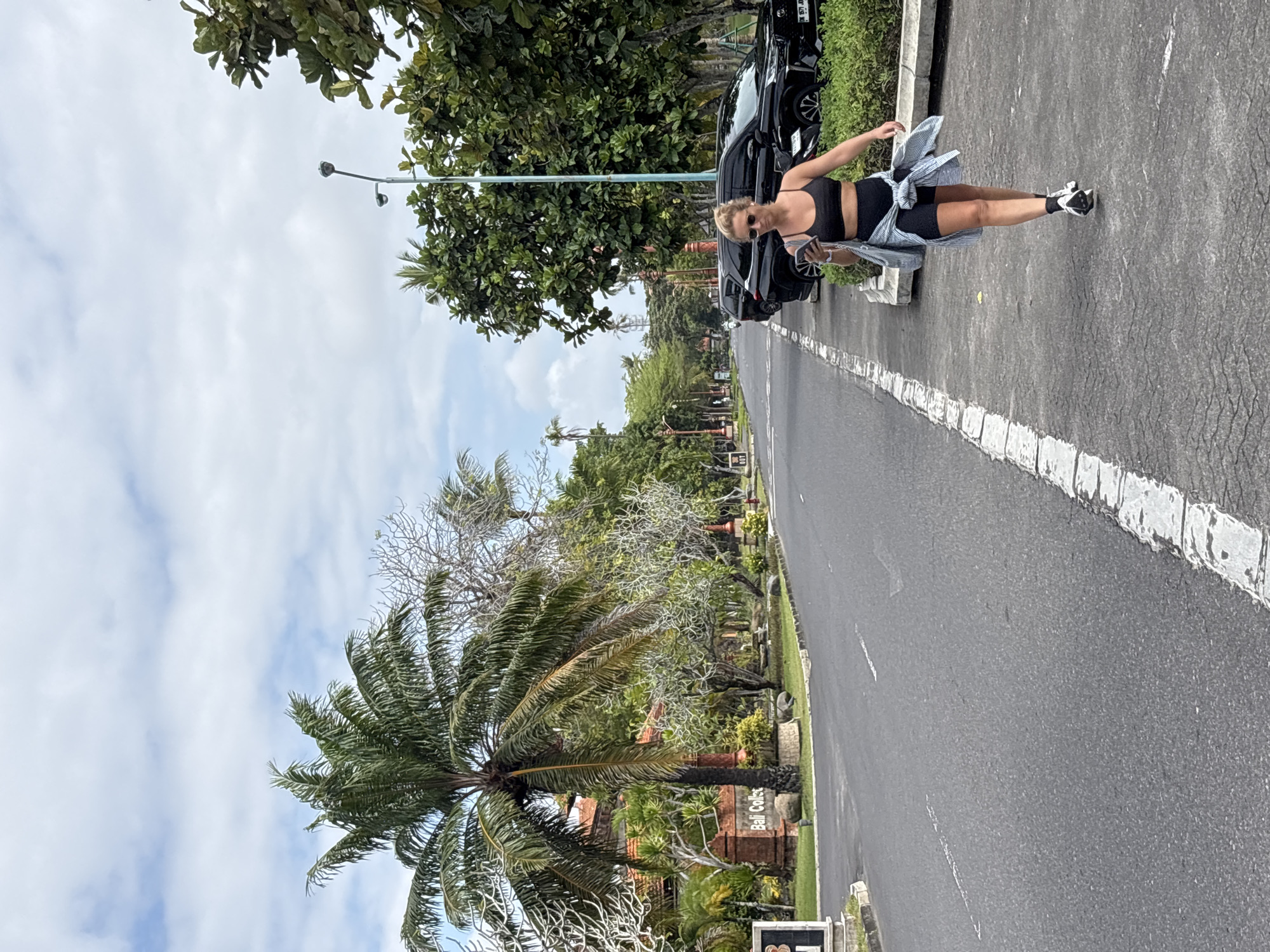 In Kuta Selatan, Indonesia, a blonde woman in athletic attire, a black sports bra and black shorts, walks along a paved sidewalk next to a road. She holds a phone in her right hand and has a light blue and white striped shirt tied around her waist. Her hair is pulled back in a ponytail, and she wears sunglasses. The sidewalk has white painted lines and is bordered by a curb and a line of green bushes. The road is asphalt with white lane markings. To the left of the road, there is a sign that reads "Bali Collection" on a brick structure, with palm trees and lush green vegetation behind it, including some buildings with orange-tiled roofs. To the right of the road and behind the woman, several black SUVs are parked. The sky is partly cloudy with a bright, hazy sun.