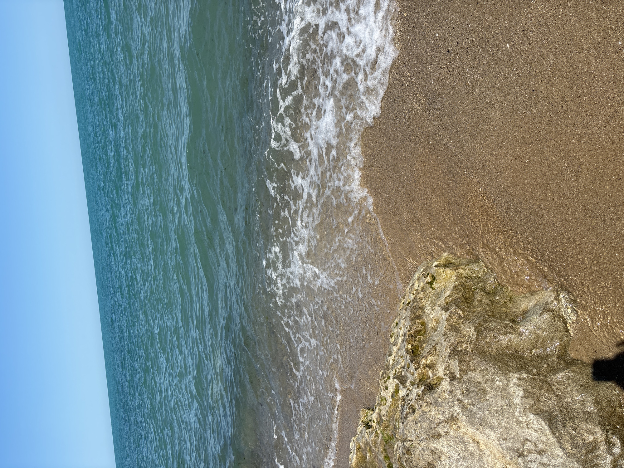 The image depicts a clear day at the beach with a calm sea and a sandy shore. In the foreground, a large, textured rock sits partially submerged in shallow water. The water gently laps at the sand and the rock, creating white foam. The sand is light brown and appears to be mixed with small pebbles. The sea is a vibrant turquoise color and stretches to the horizon under a bright blue sky. The scene suggests a peaceful coastal environment, likely a public beach area in Varna, Bulgaria, given the context. A shadow of a person, presumably the photographer, is visible in the lower left corner of the image.