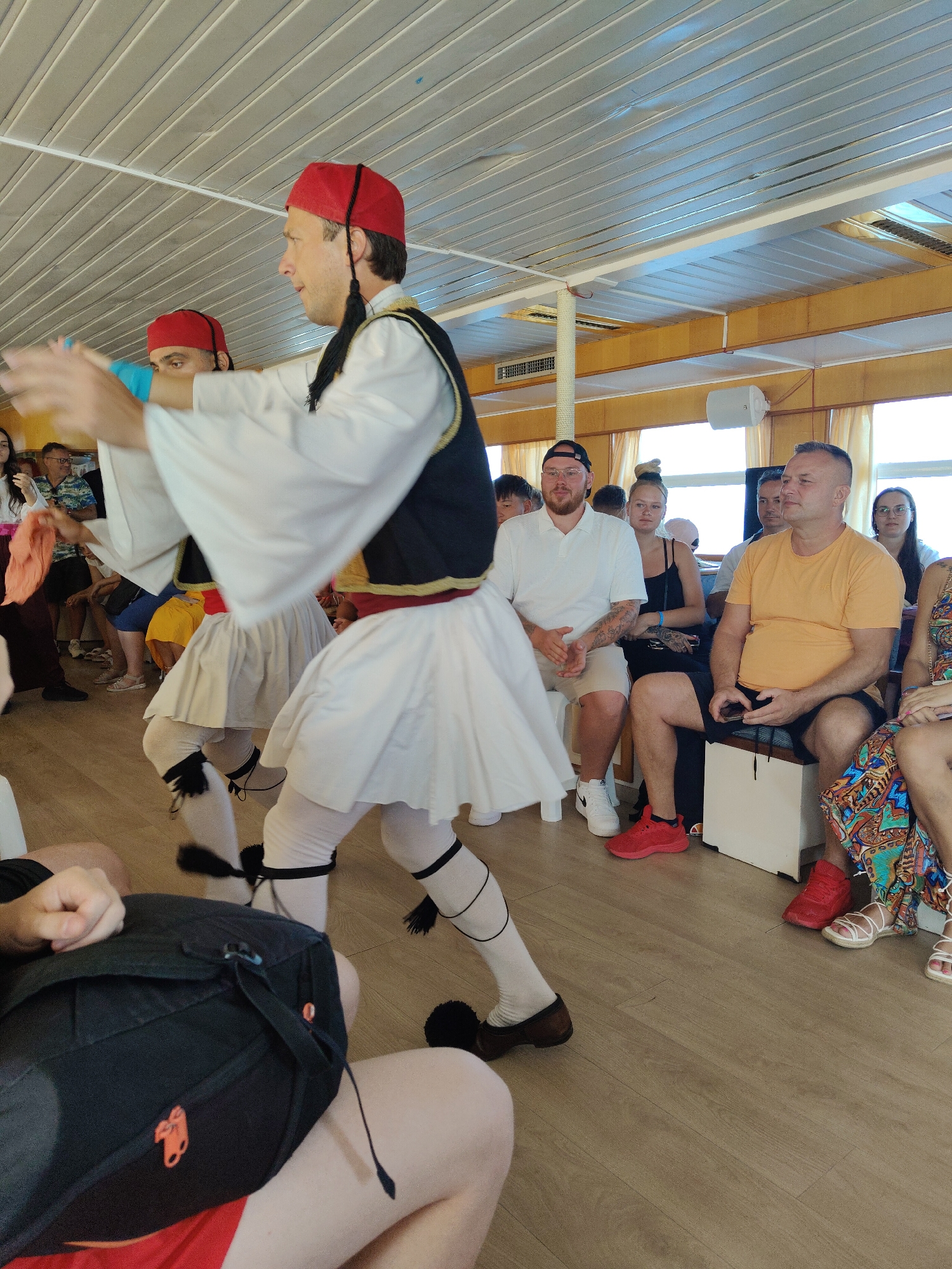 In an interior space with a slatted ceiling and light wooden floor, two men in traditional Greek attire are performing a dance. They wear red fez hats with black tassels, white shirts with voluminous sleeves, dark vests with gold trim, and white pleated skirts over light-colored tights. Their legs are adorned with black ribbons and pom-poms. The men are positioned towards the left and center of the frame, actively engaged in their dance.
Seated on the right and in the background are several spectators. A man in an orange t-shirt and dark shorts is holding a smartphone. Next to him, a woman with dark hair wears a black tank top and black shorts. Another man with a beard is seated in a white polo shirt and beige shorts, wearing red sneakers. Further back, a woman in a black tank top is visible. The overall impression is that of a cultural performance or demonstration taking place indoors. The scene appears to be on a boat, given the slatted ceiling and the presence of a railing visible in the background. The location is unspecified, but the attire and performance suggest Greece.