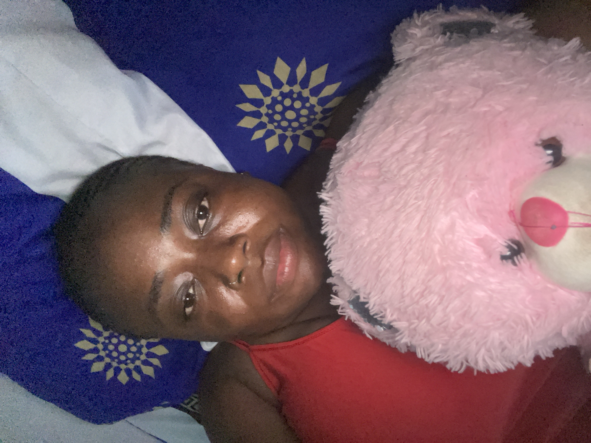 The image shows a person with dark skin and short dark hair, wearing a red tank top, lying on their back. Their head is turned slightly towards the viewer, and they are looking directly into the camera with their eyes open and lips slightly parted. The person's skin exhibits a natural sheen across the forehead, nose, and cheeks. To their left, a large, fluffy, light pink teddy bear with dark eyes and a pink nose rests beside them. The person and the teddy bear are situated on a bed with bedding consisting of a blue fabric adorned with a repetitive yellow-gold geometric pattern, resembling a stylized sun or flower, and a plain white sheet or pillowcase. The lighting suggests an indoor setting. There is no discernible information within the image to indicate the specific location in the city of Abuja, Nigeria.