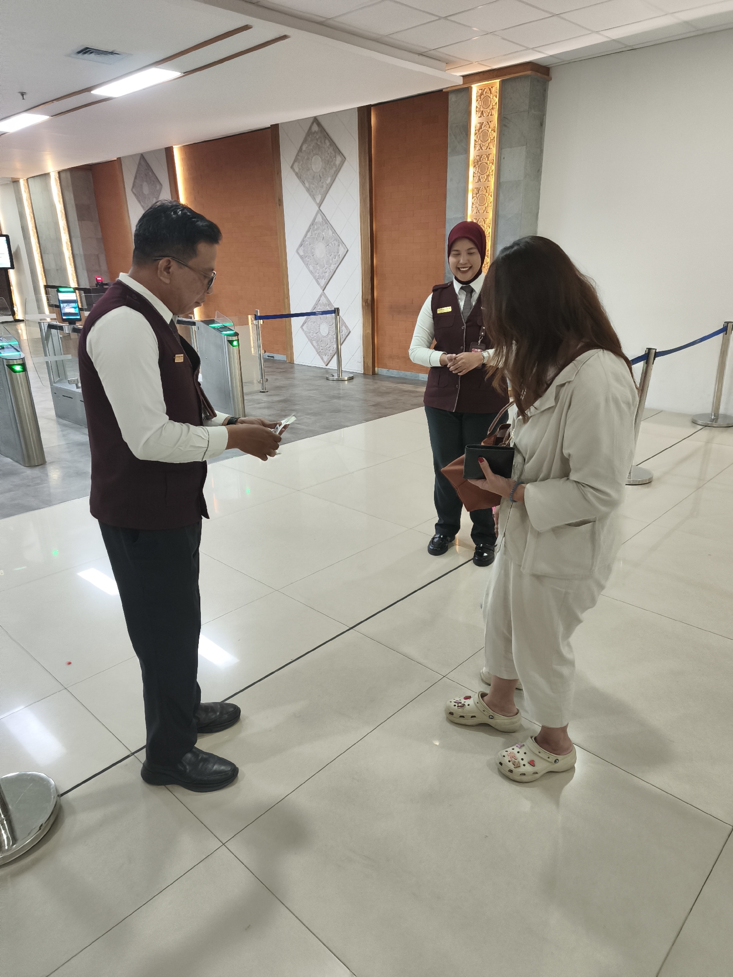 An airport security checkpoint in Kuta, Indonesia features three individuals. A male security officer in a maroon vest and white shirt stands to the left, examining a mobile phone. To the right, a female security officer in a maroon vest and hijab smiles and stands behind a woman in a beige jumpsuit who is holding a wallet and a handbag. Both security officers are wearing name tags. The environment is a modern airport terminal with tiled flooring, automated gates, and decorative wall panels.
