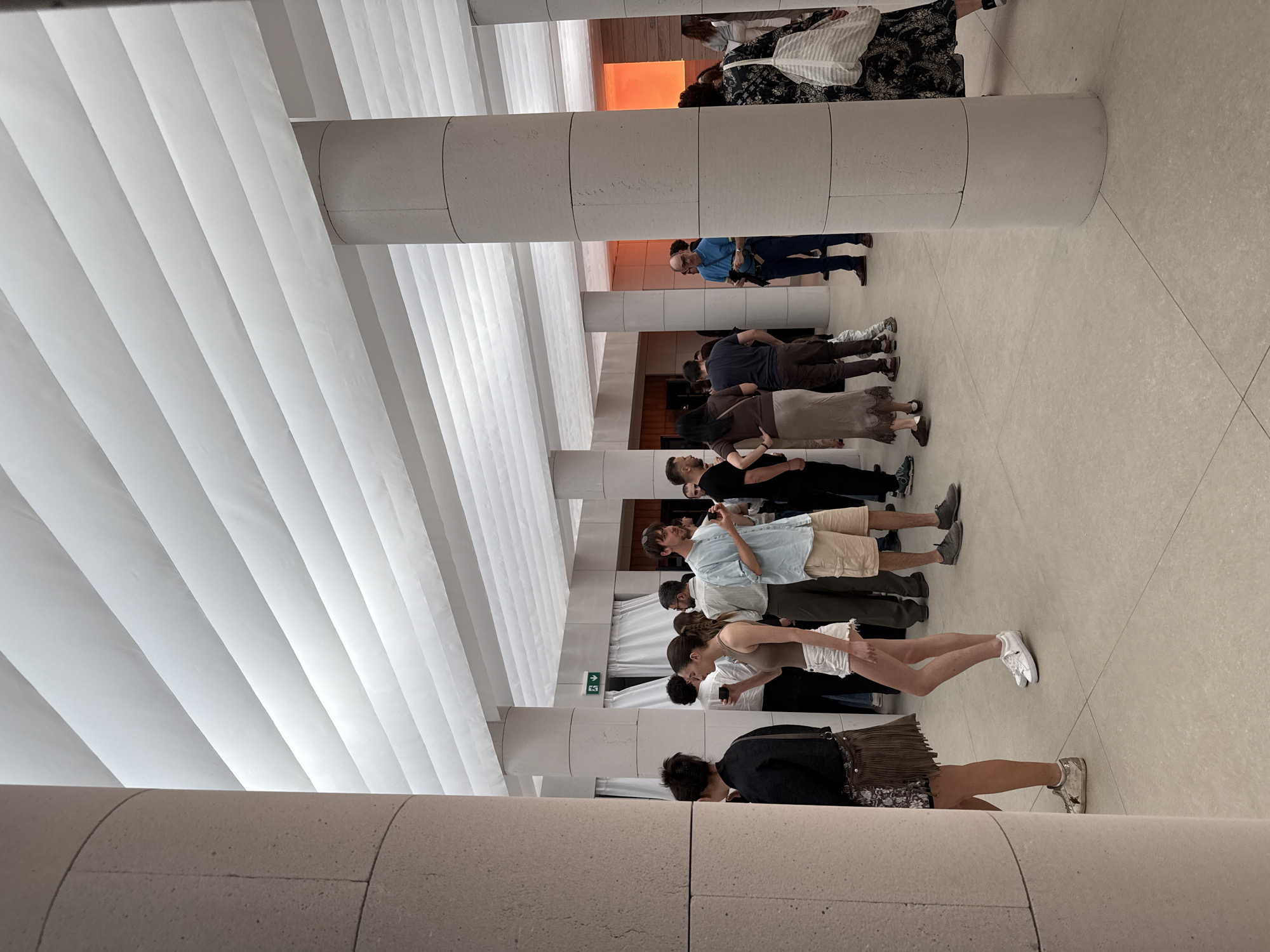 A group of people are standing in a line inside a building. The building has a modern design with a white, ribbed ceiling and cylindrical pillars. The floor is tiled. The people are diverse in appearance and clothing. Some are looking at their phones. The location is likely a museum in Milan, Italy.