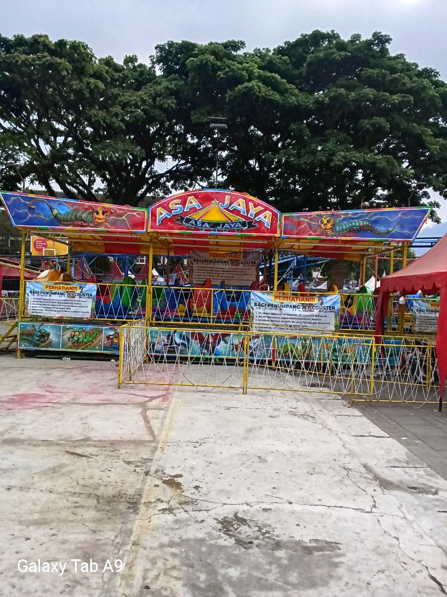 A colorful, small amusement park ride, likely a kiddie coaster, is set up outdoors. The ride features cartoon caterpillars and a sign with "ASA JAYA" prominently displayed in red and yellow. Banners with Indonesian text, likely safety instructions, are attached to the ride structure. Yellow and white temporary fencing surrounds the attraction. A large, mature tree with dense green foliage dominates the background, suggesting a park or public space. The ground is paved concrete, showing signs of wear and tear, with some puddles and stains. The overall impression is that of a temporary fairground setup. The image is watermarked with "Galaxy Tab A9" at the bottom left.