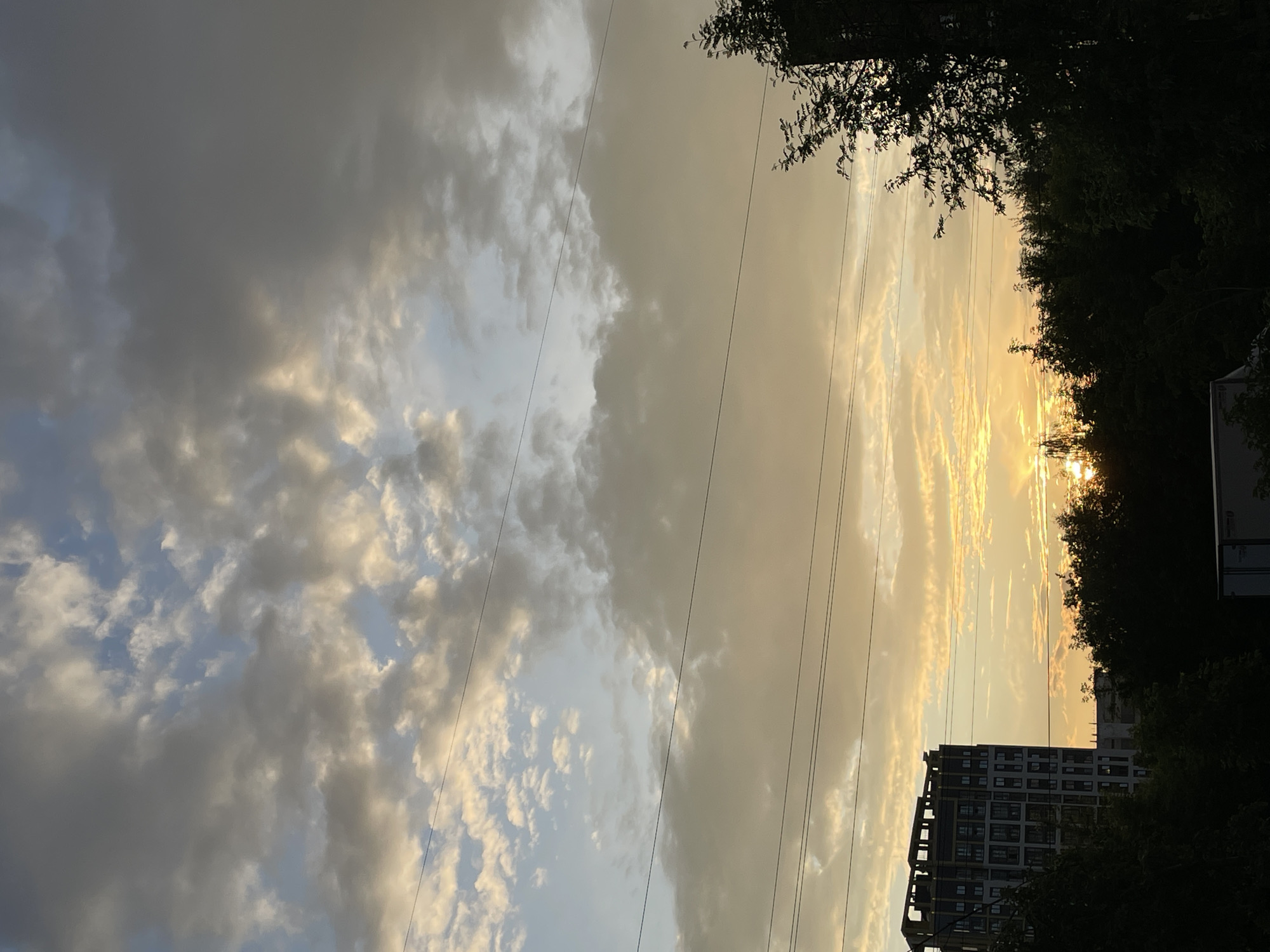 The image shows a city skyline with a dramatic sunset. In the foreground, dark green trees line the bottom of the frame, with a partially visible white truck on the lower right. To the left, a tall, modern building with a dark facade and many windows stands prominently. The sky is filled with a mix of dark gray and lighter, yellowish clouds, with the sun peeking through the horizon, casting a warm glow. Several power lines are stretched across the sky horizontally. The scene appears to be captured from a slightly elevated viewpoint, looking towards the west as the sun sets.