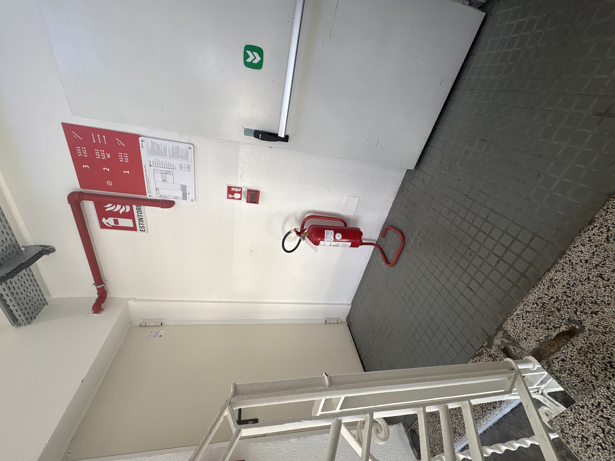 This image captures an indoor stairwell landing, likely within a commercial or public building in Milan, Italy, as indicated by the Italian text on various signs. The shot is taken from an elevated, tilted perspective, looking down towards a lower floor or landing.
The primary subjects include a white emergency exit door, a fire extinguisher, and various safety signs. The white door, equipped with a silver panic bar and a prominent green exit sign featuring chevron arrows, leads outwards. Adjacent to the door on the white wall, a red fire alarm call point is visible. On the floor below, a red portable fire extinguisher lies on its side.
To the left of the door, a red informational sign is mounted on the wall. It details floor numbers and corresponding room numbers: "3 S211 S212", "2 S211 S212 WC", and "1 S111 S112," with wavy lines symbolizing staircases. Next to this, an emergency evacuation plan is displayed, titled "Piano di Evacuazione / Evacuation Plan." Below these signs, a red fire suppression pipe runs along the wall, with a small red sign featuring a fire extinguisher icon and the word "ESTINTORE" (Italian for extinguisher).
The flooring varies, with a grey square-patterned tile area near the exit door transitioning to a darker, speckled terrazzo-like material on the lower landing and visible stairs. A white, ornate metal railing for the staircase extends into the lower right of the frame. On the far left, part of a metallic cable tray system is visible. The walls are painted white, and the area appears well-lit, suggesting either natural light from a nearby source or artificial lighting. No people are present in the scene, which appears static and functional.