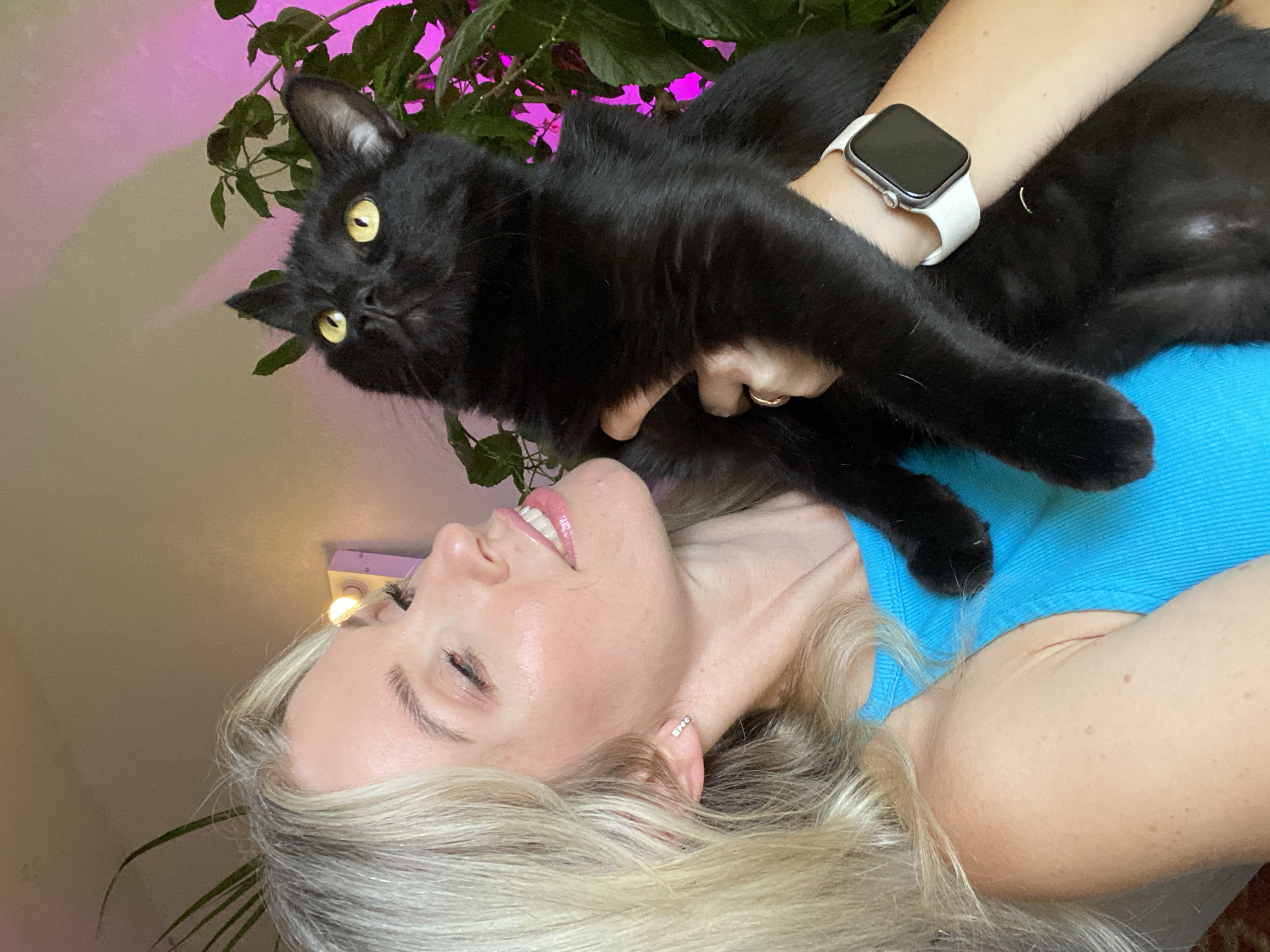 A blonde woman in a blue tank top holds a black cat. The cat is looking upwards, and its paws are visible. The woman is looking at the cat. A pink light illuminates a plant in the background.