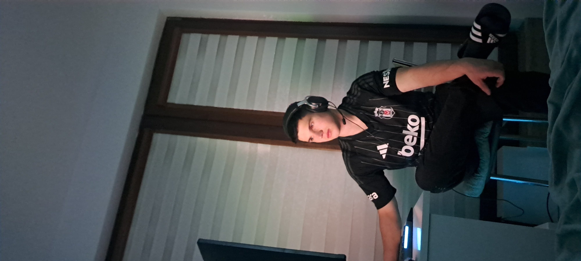A young man sits in a chair, wearing a black and white soccer jersey with headphones on. His left leg is raised, with an Adidas sandal on his foot. He is in front of a computer monitor, with closed white blinds behind him. The image does not contain enough information to determine if it is Burgas, Bulgaria.
