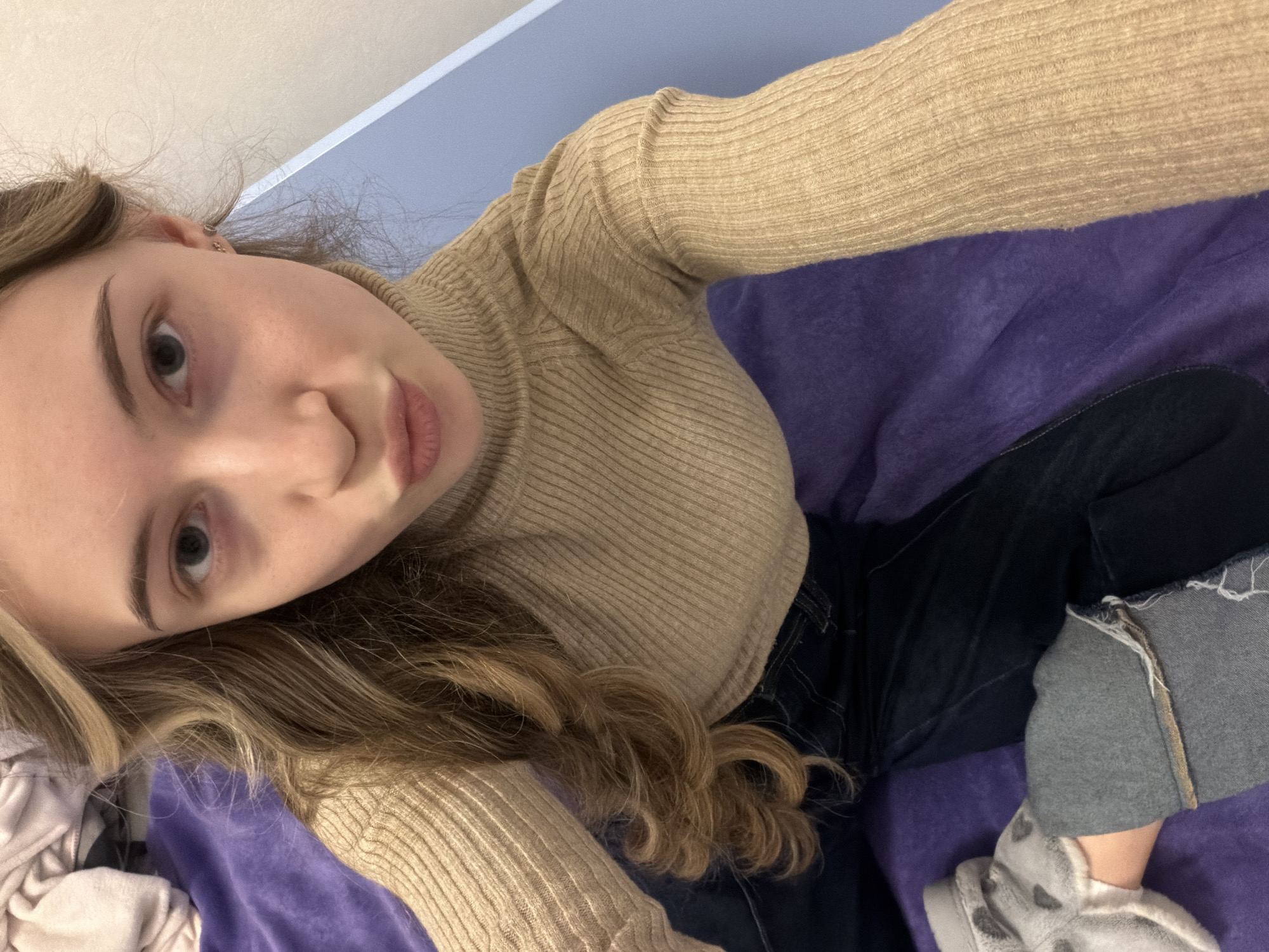 A selfie taken from an overhead angle shows a young woman indoors. She is lying on a purple surface, possibly a bed or couch. She wears a fitted, tan-colored ribbed turtleneck sweater and dark blue jeans. Part of her leg is visible, covered by a gray fabric or garment and a gray and white patterned shoe. The background includes a light blue wall or surface. There is no information in the image to determine the location in the city of Plovdiv, Bulgaria.