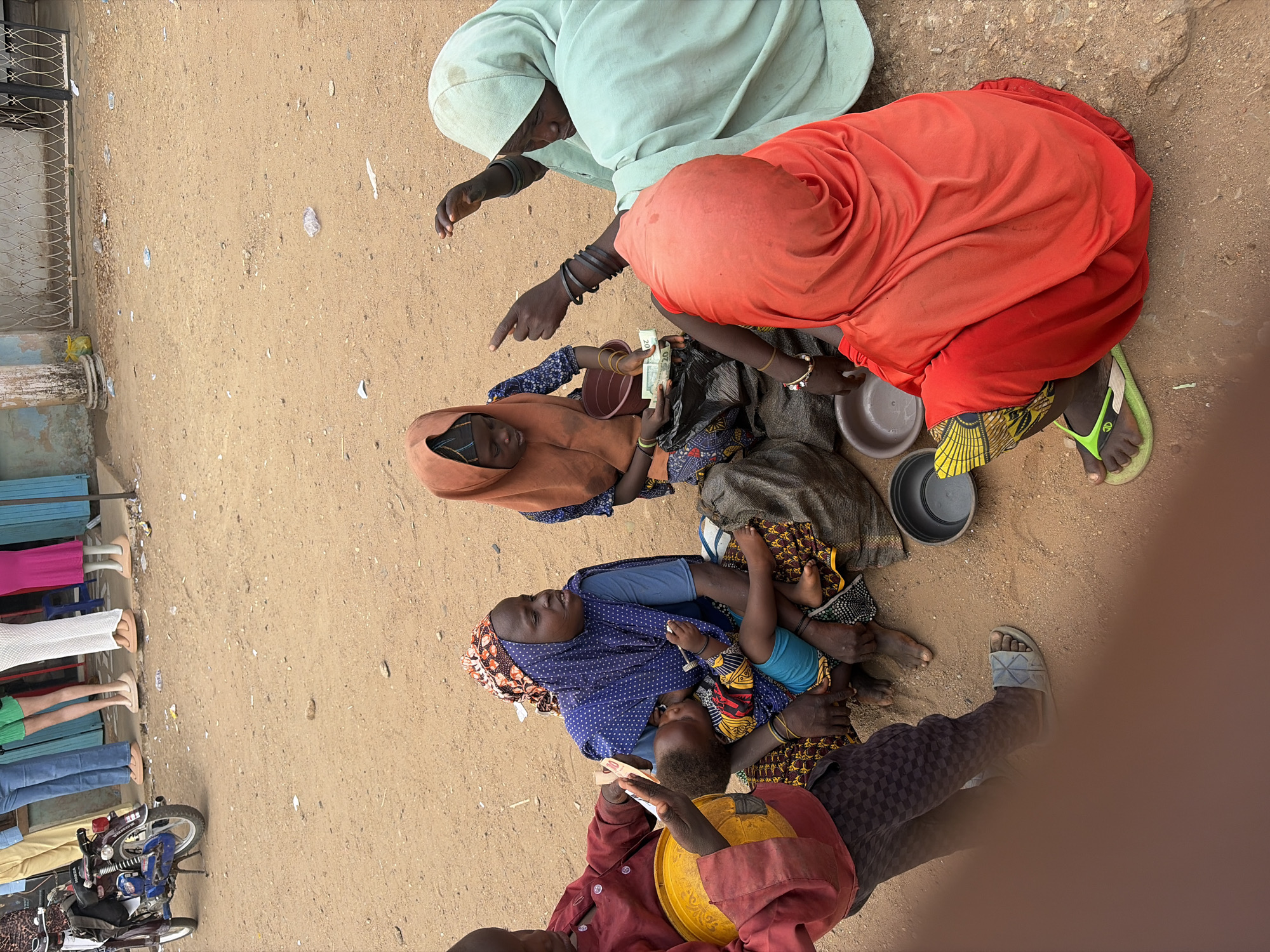 This media file presents an overhead view of an outdoor scene in Jos, Nigeria, characterized by an unpaved, sandy-brown ground under bright daylight.
In the center of the frame, a group of women and children are seated directly on the ground. One woman, covered in a light green headscarf and garment, extends her arm with a pointing finger. Adjacent to her, a woman in a vibrant orange headscarf and flowing dress is also seated. Below them, a third woman, wearing a brown headscarf and garment, is seen holding several banknotes, one of which clearly shows the number "20." Near her is a small, empty, reddish-brown bowl. Her wrists are adorned with dark bangles.
Further towards the lower part of the image, another woman in a blue patterned headscarf and garment cradles a baby or toddler, dressed in a blue top and patterned shorts. A young child or person in a red top and dark checkered pants sits nearby, holding a yellow bowl. Several small, grey plastic bowls are scattered on the ground around the seated individuals. Footwear, including flip-flops, is visible on the feet of some individuals.
To the left of the scene, a blue wall with a black metal fence or grill runs parallel to the ground. Below this, a line of clothing mannequins, displaying only legs and feet clad in various trousers (pink, white shorts, green shorts, blue jeans, and tan pants), stand upright. A blue and silver "HONDA" motorcycle is parked nearby. The ground also shows scattered small pieces of litter and a yellow plastic bag.
The overall scene depicts a bustling street or marketplace environment, with interactions possibly related to informal trade or social gathering, under the open sky.