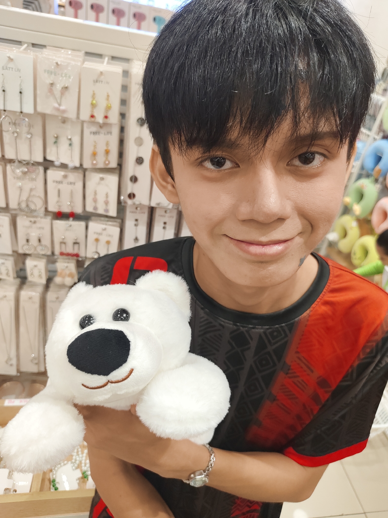 A young person with dark hair and eyes is holding a white stuffed bear in their left arm, looking directly at the camera with a slight smile. They are wearing a black t-shirt with red accents and geometric patterns. Behind the person, a display of various earrings and necklaces is visible, suggesting a retail environment. The scene appears to be inside a store in Davao City, Philippines, possibly a boutique or a department store. The lighting is bright and even, illuminating the subject and the merchandise. The background is slightly blurred, drawing focus to the person and the bear.