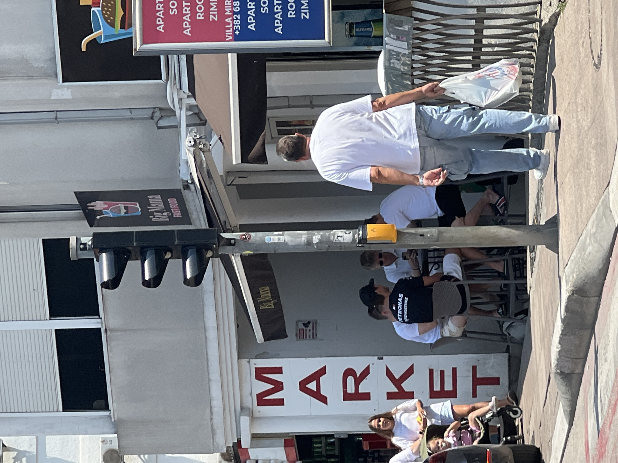 The image depicts a street scene in Budva, Montenegro, on a sunny day. In the foreground, a man wearing a white t-shirt and light blue jeans walks away from the camera, carrying a white plastic bag in his right hand. He is on a sidewalk next to a street with a concrete curb. To his left, a traffic light is visible. Behind him, a building houses a "MARKET" store with large red letters on a white background. Several people are seated at tables outside the building, appearing to be at a cafe or restaurant with an awning labeled "Big Mama FAST FOOD". A young woman stands near the MARKET sign holding a baby in a stroller. Further to the right, a red and blue sign is affixed to the building, advertising "APARTMENTS" and providing a phone number. Various details of the urban environment, such as windows, shutters, and a metal fence, are also present.