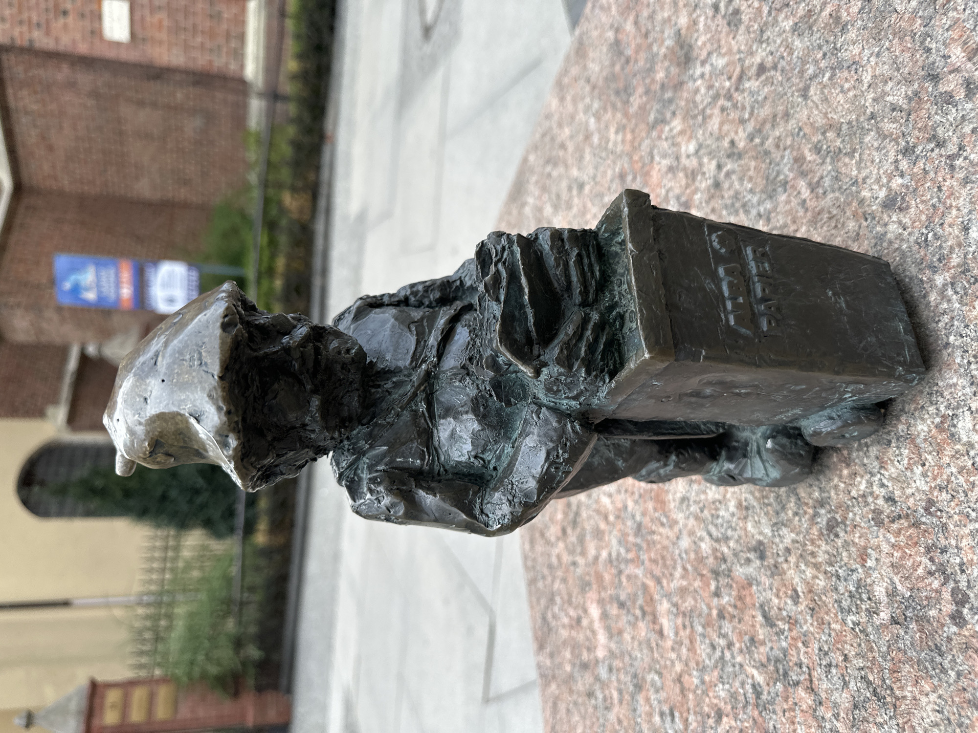 The image is a close-up of the bronze statue of a dwarf holding a ledger or tablet. The dwarf statue sits atop a mottled granite or similar stone base. The backdrop is a blurred urban scene, suggesting a building of brick with signage and possibly foliage. The word "ALBA" is legible on the tablet the dwarf holds. Based on the style and information, the statue likely is part of the collection of Wrocław dwarfs found throughout the city of Wrocław, Poland.