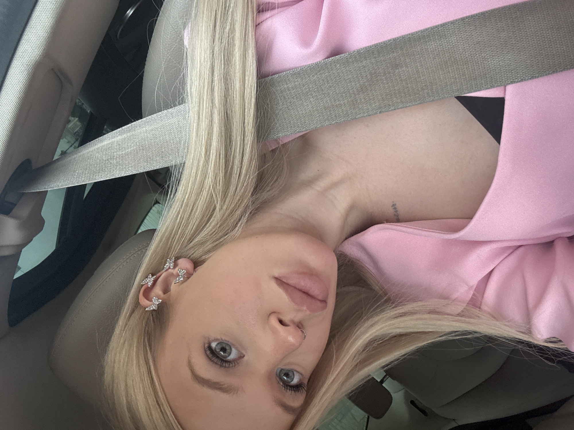 A young woman with blonde hair and blue eyes is in the driver's seat of a car. She is wearing a pink jacket and a black bra. She has several butterfly-shaped earrings in her left ear and a nose ring. A seatbelt crosses her chest. Her hair is long and blonde. The background of the image shows the interior of a car, including a seatbelt and the car's roof. The image appears to have been taken during the daytime. The specific location in Saint Petersburg, Russia is not discernible from the image.