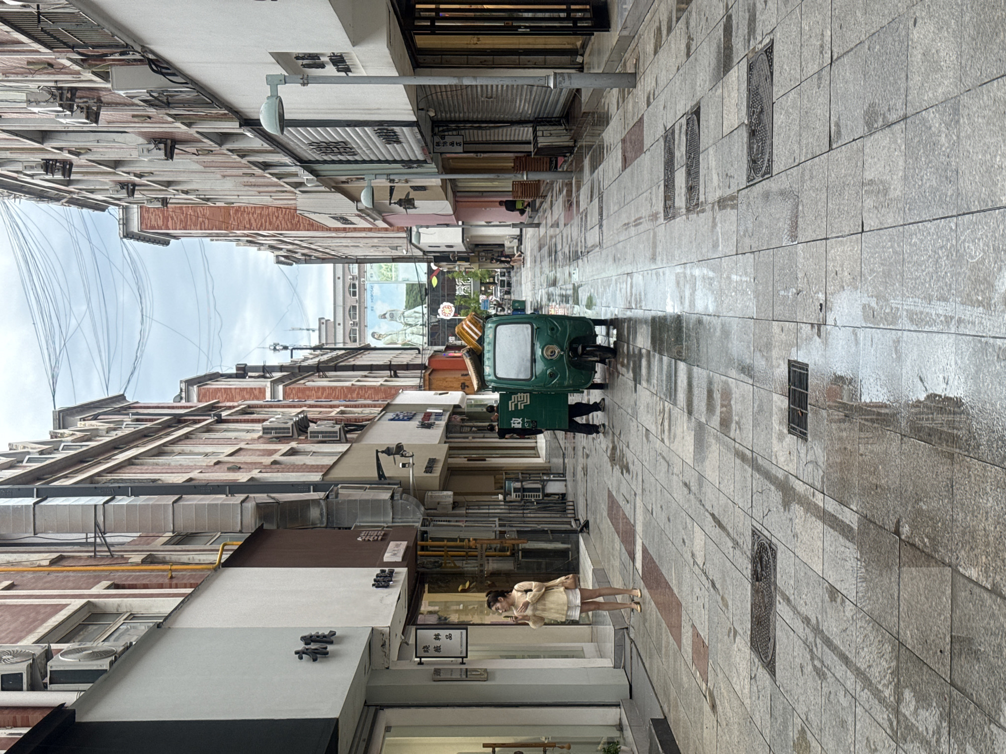 The image shows a narrow street in Qinhuangdao, China, with buildings lining both sides. The street is paved with large, grey tiles and appears wet, suggesting recent rain. There is a green, three-wheeled vehicle parked in the middle of the street, facing away from the camera. A person wearing a light-colored top and white shorts stands on the left side of the street, looking to their left. Another person wearing dark clothing and a green vest with a white box design on their back is standing near the green vehicle, also facing away from the camera. Several shop fronts are visible on the buildings, some with their shutters down. The sky is overcast.