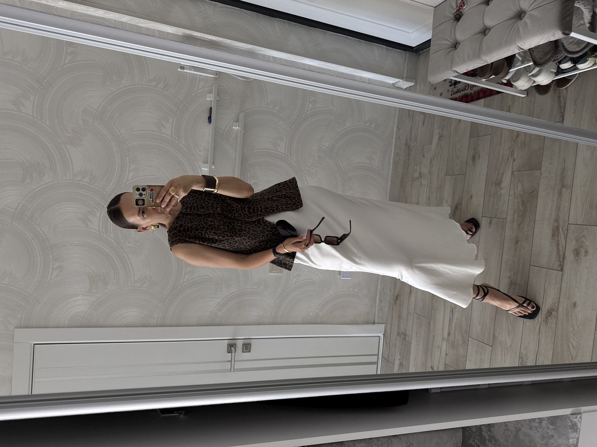 In a room with light wood-paneled flooring and textured white wallpaper, a woman stands in front of a full-length mirror, taking a selfie. She is wearing a sleeveless, leopard-print vest over a flowing, ivory satin maxi skirt. Her hair is pulled back into a bun, and she is adorned with large, gold hoop earrings, a gold cuff bracelet, a gold ring with a green stone, and a black wristwatch. She holds a pair of dark sunglasses and a black clutch in her right hand. Her black strappy sandals with ankle ties are visible on her feet. The room appears to be an interior space, possibly a home, with a white door with silver hardware visible in the reflection to the left. To the right, a grey tufted bench with shelves holding shoes is partially visible. The scene does not contain any specific indicators to place it in Sochi, Russia.