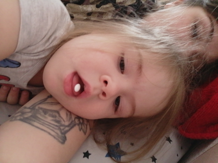 The scene is an indoor setting, likely a bedroom, located in the City of Chaikovsky, Russia. A young child, with light-colored hair, is depicted lying down, angled towards the viewer. A small, white, circular pill is visible on the child's extended tongue. The child wears a light-colored t-shirt with subtle, partially obscured patterns. To the lower left of the frame, an adult's bare arm, adorned with a prominent black ink tattoo depicting an ornate crest-like design, is visible. This arm likely belongs to the individual capturing the image. In the upper right portion of the background, a partial reflection of an adult's face and hair is discernible. The immediate surface beneath the child is a white textile with a pattern of dark-colored stars. Additional background elements include red knitted fabric and a darker, possibly camouflage-patterned textile.