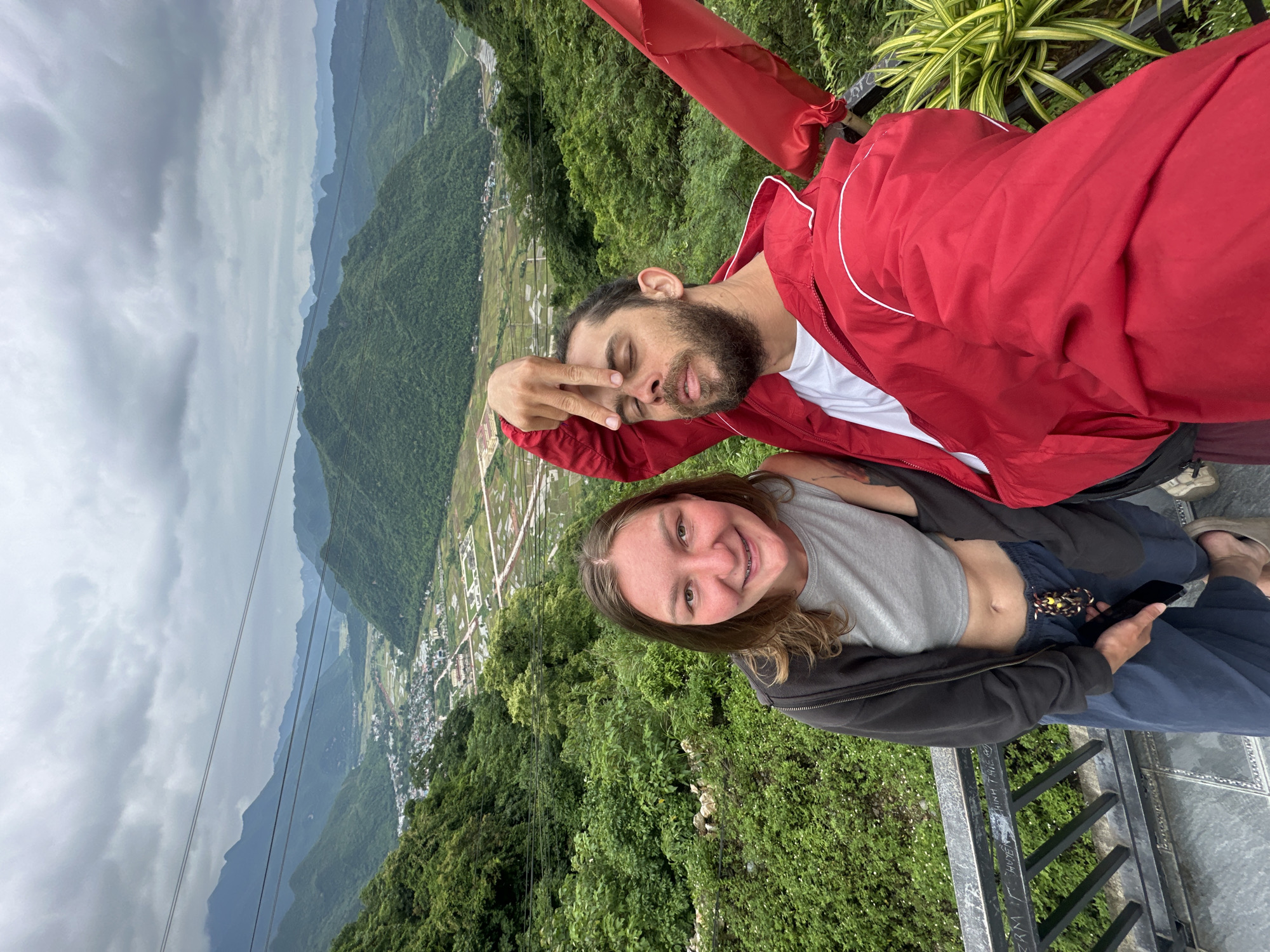 Two people, a woman and a man, are taking a selfie on a balcony overlooking a valley in Mai Chau, Vietnam. The woman is in the foreground on the left, smiling and wearing a gray crop top, a dark gray zip-up hoodie, and dark blue pants. She has braces on her teeth. The man is on the right, wearing a white t-shirt and a bright red jacket. He is making a gesture with his hand over his eyes. Behind them, lush green mountains dominate the landscape, with a town nestled in the valley below. The sky is cloudy, suggesting a temperate climate. The balcony has a metal railing with some writing on it. There is a red flag attached to a pole on the right side of the frame.
