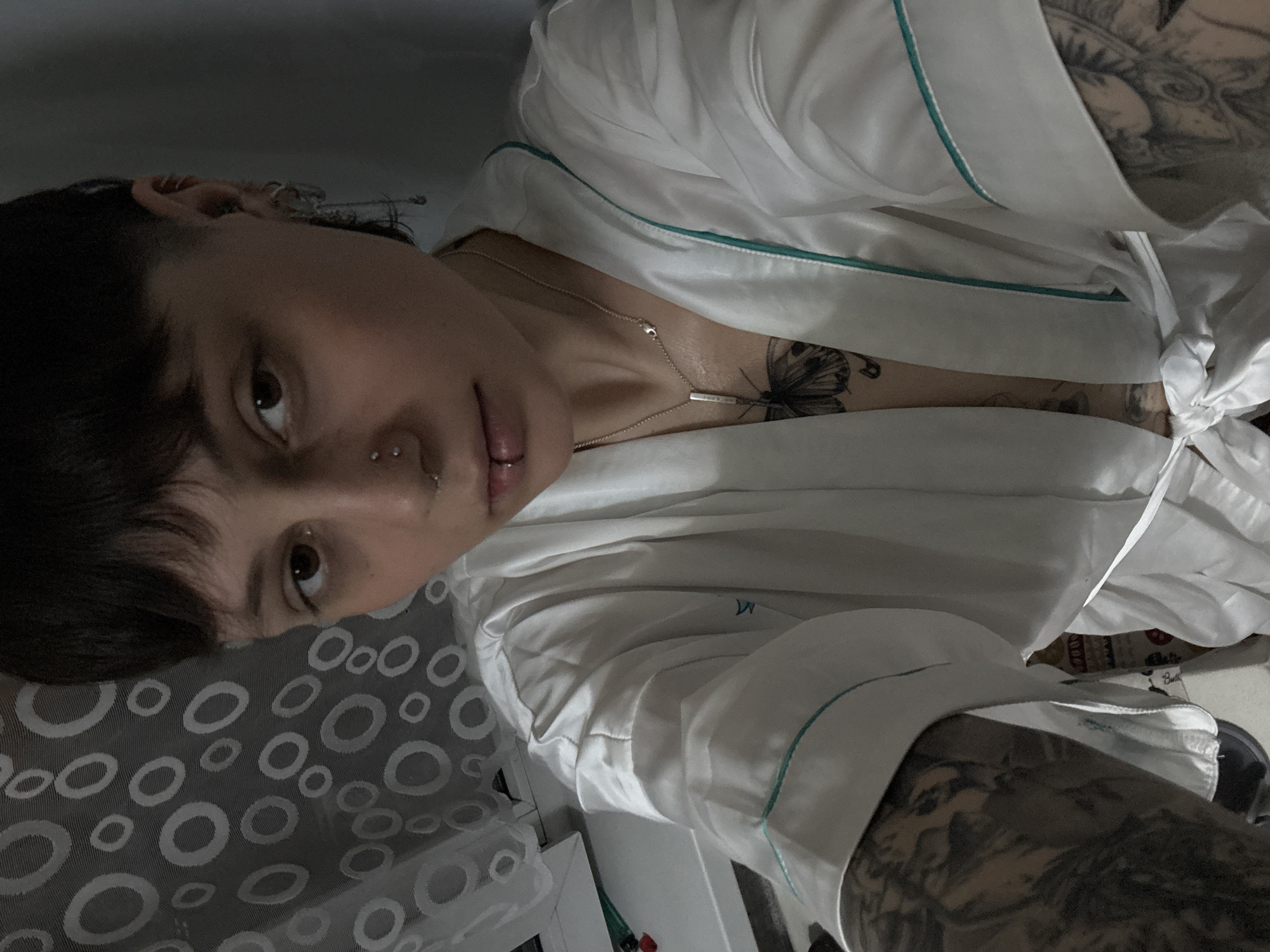 A close-up, slightly angled shot captures a person indoors. They are partially reclined on a surface covered with a patterned fabric in shades of gray and white. The person is wearing a white satin robe with teal trim. Tattoos are visible on their upper chest and arms. A silver necklace with a bar pendant adorns their neck. They have dark, short hair, and facial piercings are present. The background includes glimpses of furniture and other items. There is no evidence to support the location being Saint Petersburg, Russia.
