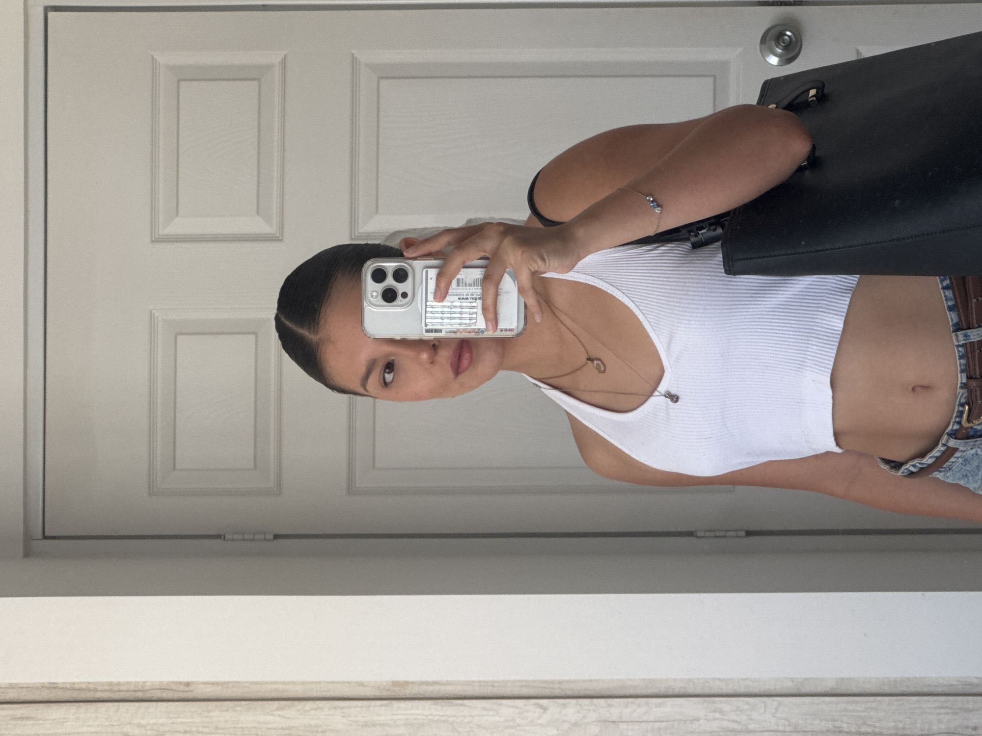 A young woman is taking a selfie in front of a white door. She is wearing a white tank top and denim shorts with a brown belt. She is holding an iPhone in her right hand, obscuring part of her face. A black bag is slung over her shoulder. She has her hair pulled back. The background is a white door with silver hardware and molding. The scene appears to be indoors, possibly in a hallway or entryway. There is no indication of San Francisco Ocotlán, Mexico.