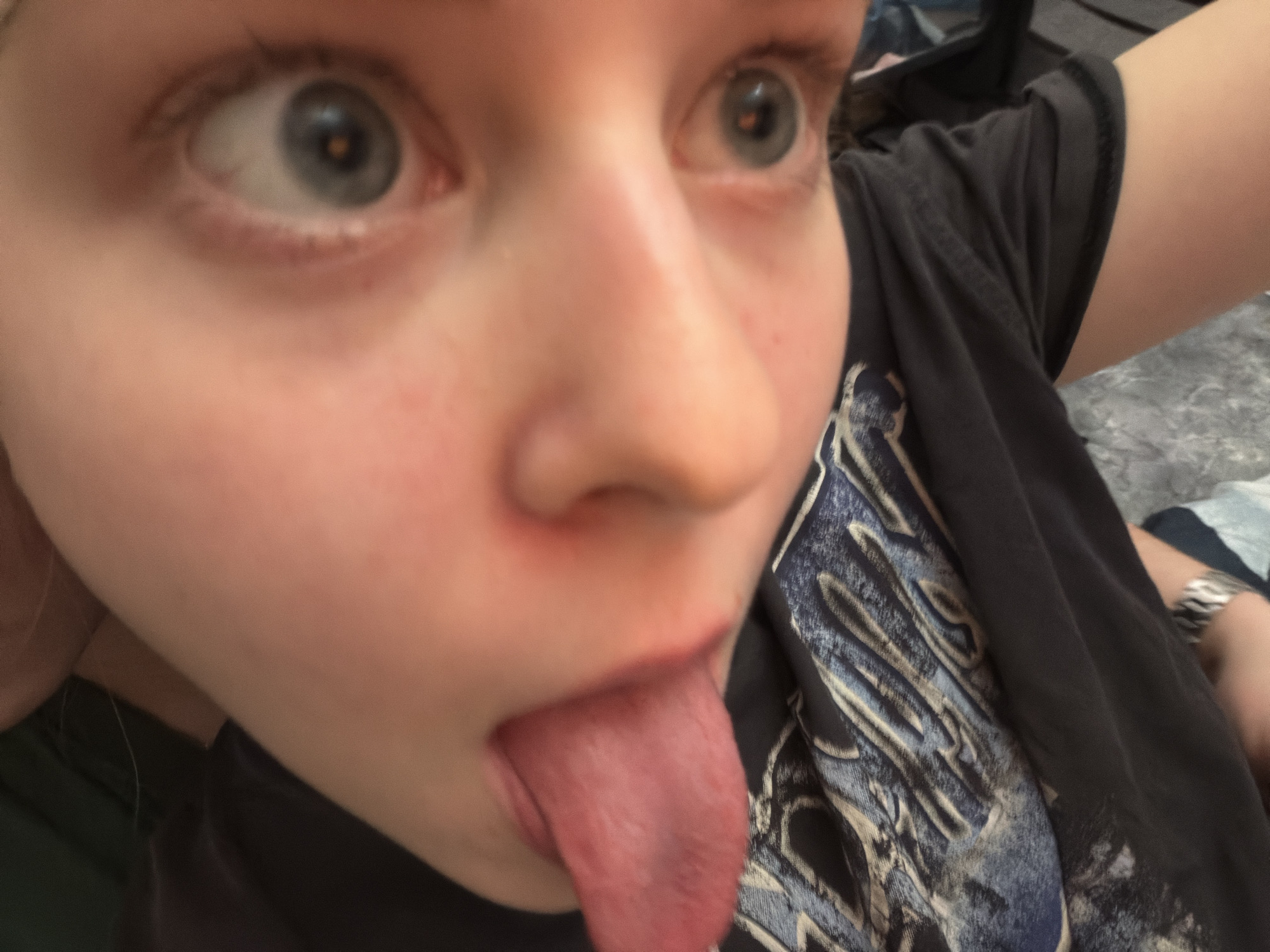 A close-up, candid photograph captures a young person making a playful expression indoors. The subject's face is positioned diagonally, with their blue-grey eyes wide open and looking upward and to the right. They are sticking out their tongue, which is fully extended. The person wears a dark-colored t-shirt featuring a prominent, light-toned graphic design on the chest. This graphic consists of stylized, textured lettering, partially visible as "IRON" within a larger, abstract pattern, resembling a band logo. A metallic-looking bracelet or watch is visible on the subject's left wrist. The background is blurred, showing indistinct grey and blue patterns, consistent with an indoor setting. The image conveys a lighthearted and silly mood. No specific time of day or weather is evident, nor are explicit visual cues to the provided location context of Perm, Russia.