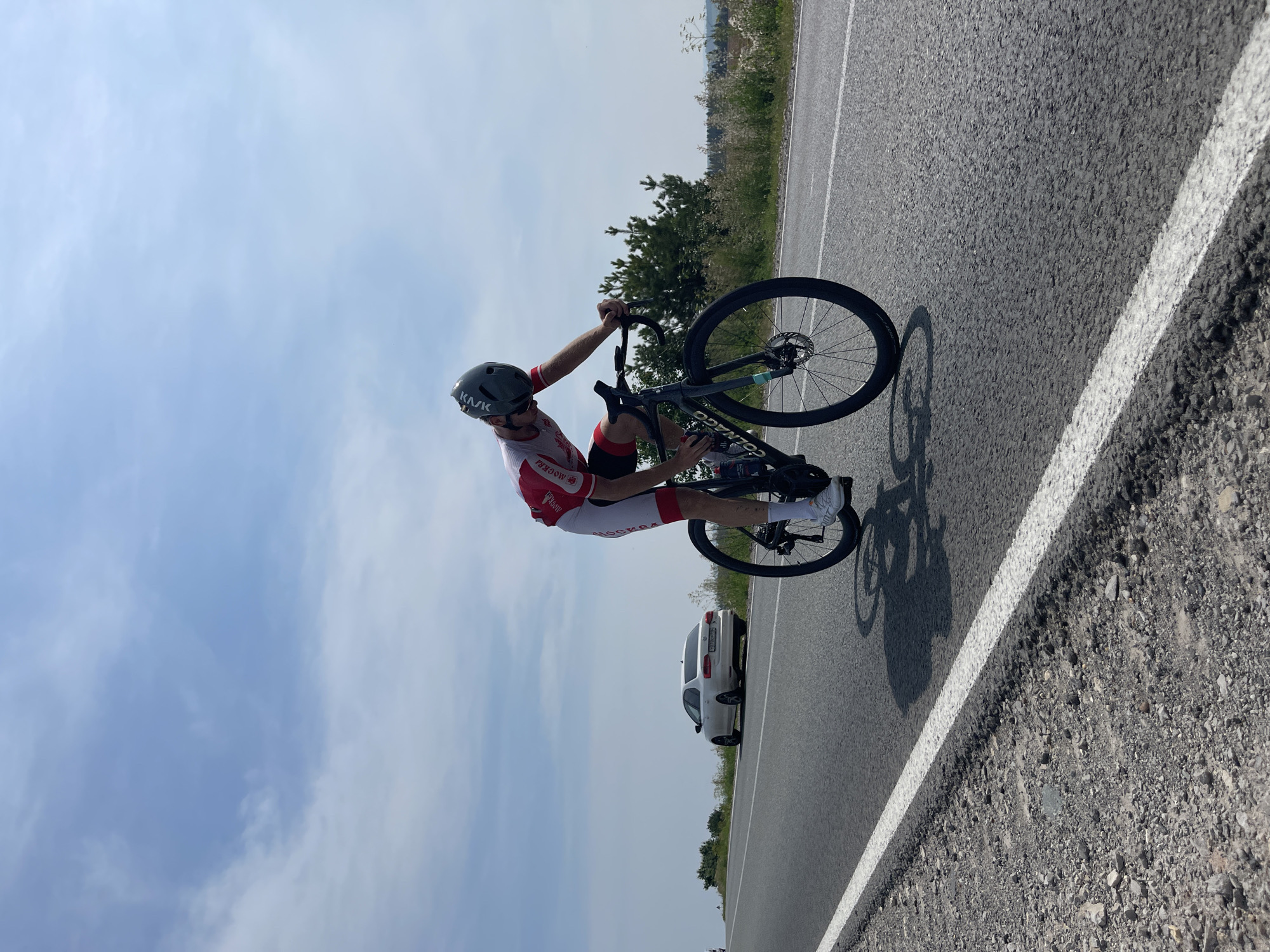 A male cyclist in a red and white racing suit and helmet is riding a black Colnago bicycle on an asphalt road. The cyclist is leaning forward, bent over the handlebars, in a racing posture. The road has a white line marking the edge. To the left of the cyclist, a white Audi sedan is parked on the side of the road, with green foliage and trees in the background. The sky is blue with scattered clouds. The location is the city of Perevoznoe, Russia.