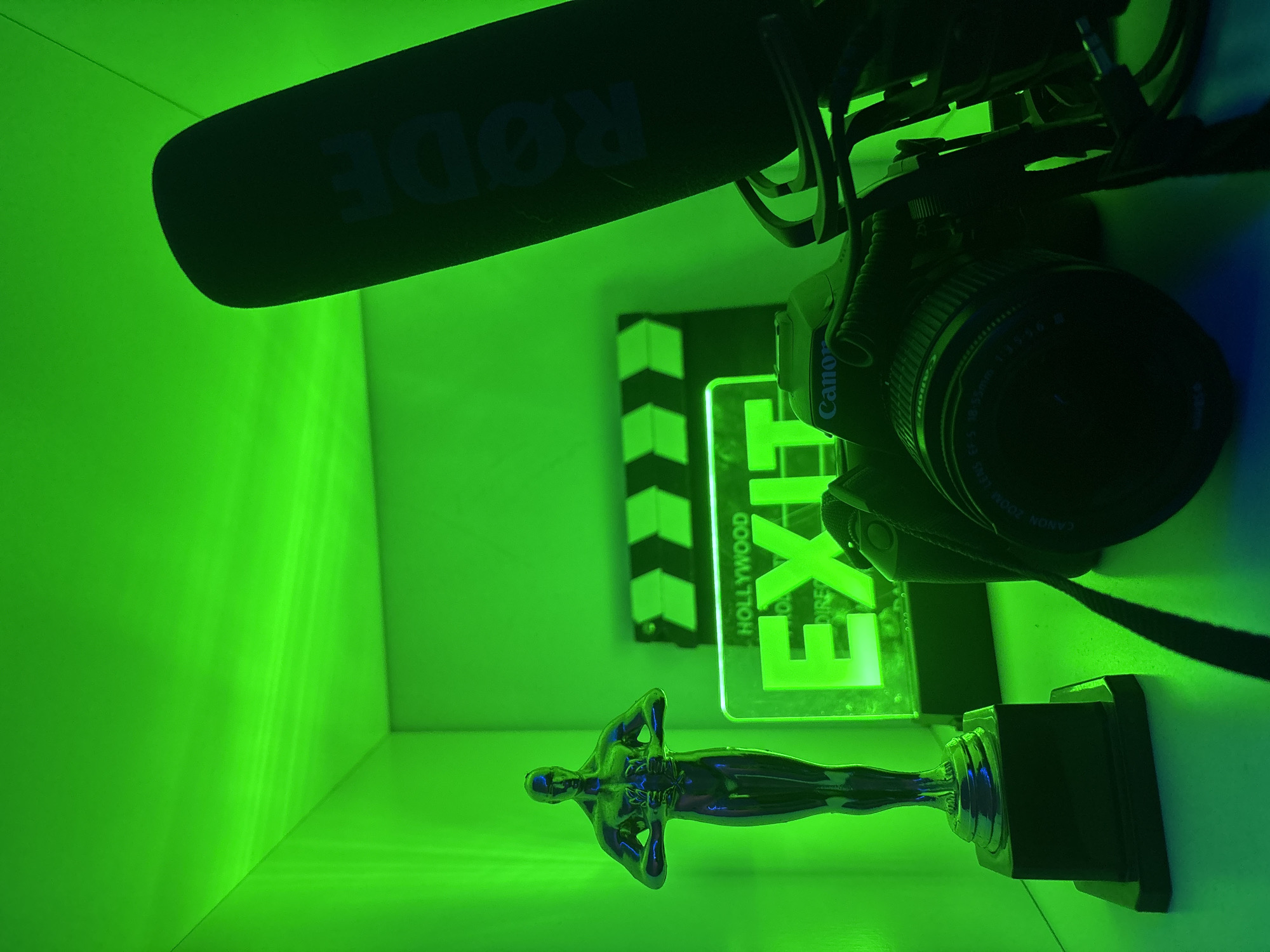 A still life composition featuring film-related objects illuminated with green light. A Rode microphone with a wind screen is at the upper left, mounted on a Canon DSLR camera with an 18-55mm lens. Below the camera, there is a film clapboard with black and white stripes and the word "HOLLYWOOD" printed on it. To the right of the clapboard is a translucent illuminated sign with the word "EXIT" in green letters. At the bottom of the composition is a silver Oscar-like statue. The objects are situated in a corner, with green light reflecting on the walls. There are no discernible people present. There is no location discernible in the city of Plovdiv, Bulgaria.