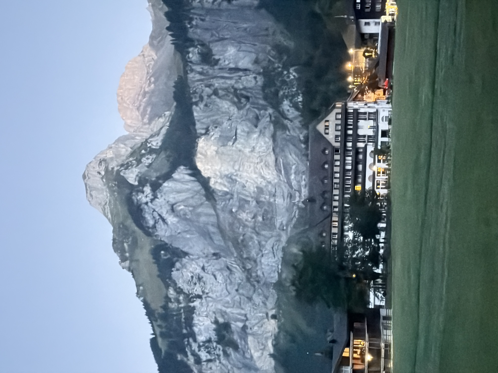 In Kandersteg, Switzerland, a large, multi-story building with a dark, sloped roof and numerous windows stands prominently. The building appears to be a hotel or a large lodge, with some windows illuminated, suggesting indoor activity. It is situated in front of a massive, imposing mountain range characterized by steep, rocky cliffs and sparse vegetation at higher elevations. The mountain faces are textured and layered, indicative of geological formations. In the foreground, a wide, well-maintained green field stretches out, suggesting a park or a sports ground. Scattered trees are visible between the field and the building, adding depth to the scene. The sky is a clear, pale blue, indicating either early morning or late evening light, with the peaks of the mountains catching a warm, golden glow. The overall atmosphere is serene and picturesque, typical of a mountain town setting.