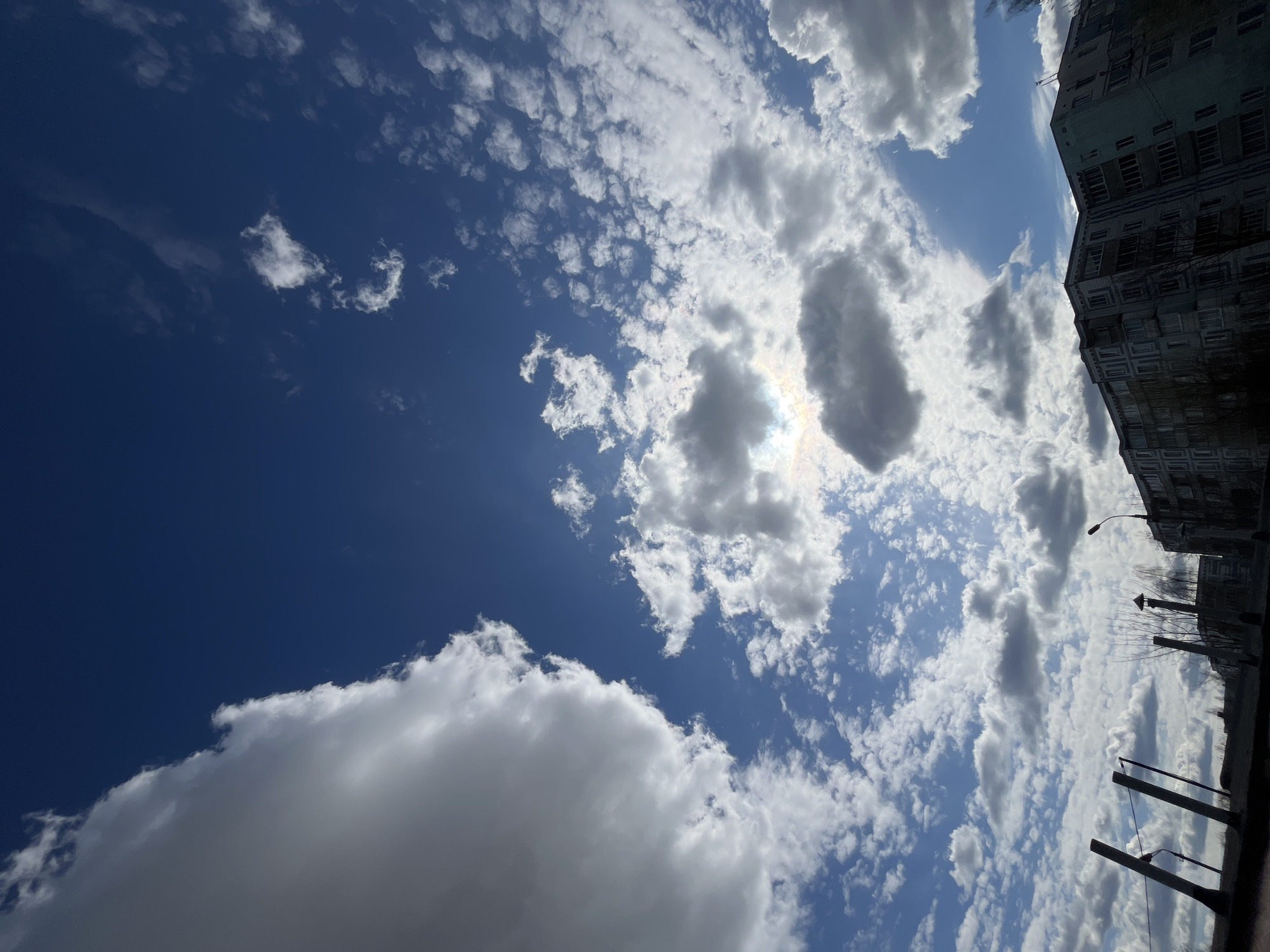 The image shows a sky view in Vysokovsk, Russia. A building with multiple windows is visible on the upper right side. The sky is blue with various cloud formations, including cumulus and cirrocumulus types. The sun is partially obscured by the clouds. Some utility poles and wires can be seen on the lower right.
