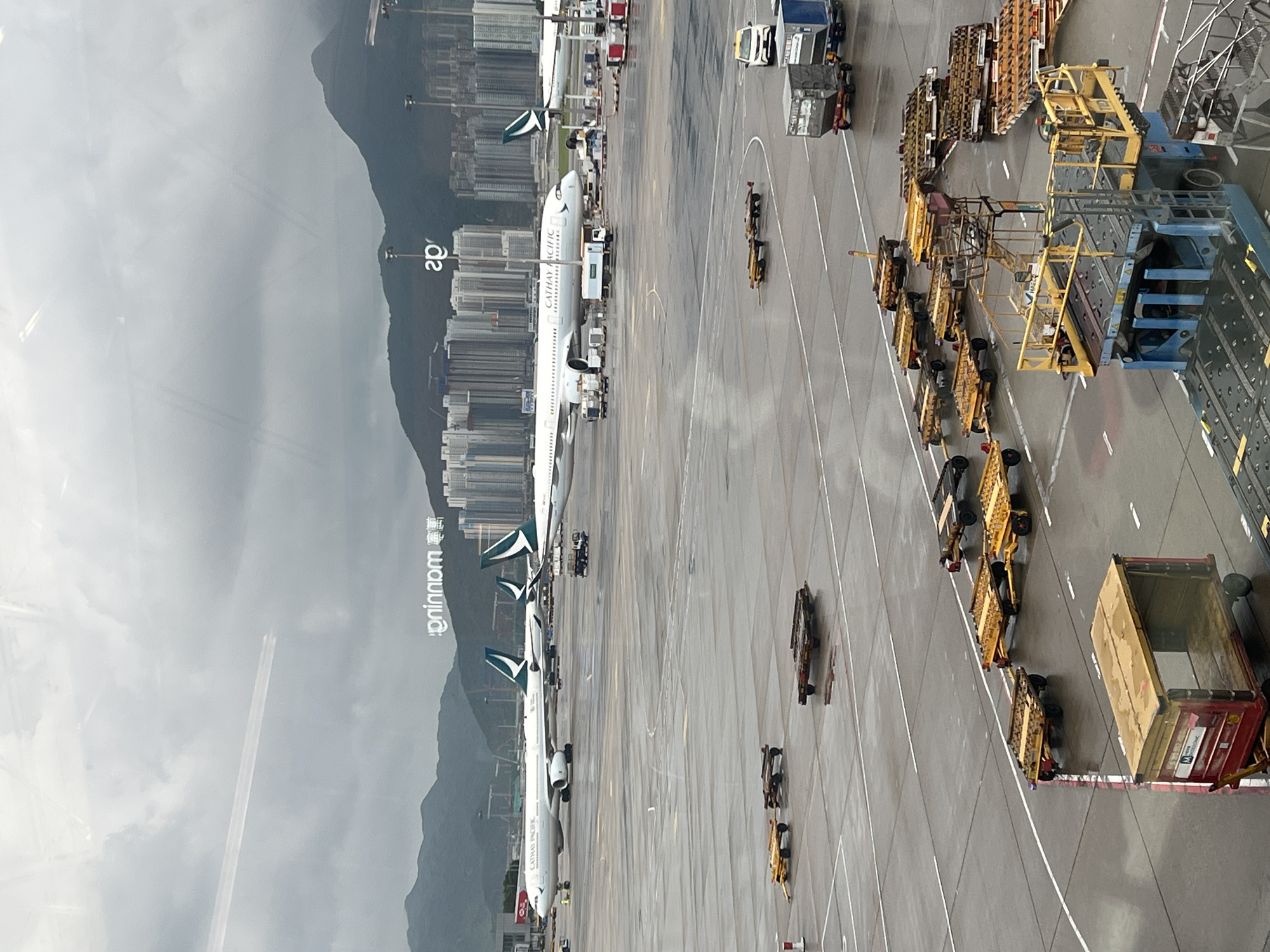 The image depicts a scene at Hong Kong International Airport (HKG) on a cloudy day. The foreground is dominated by various airport service vehicles and luggage carts on the wet tarmac. In the mid-ground, several Cathay Pacific airplanes are parked, with some appearing to be serviced. One aircraft prominently displays "CATHAY PACIFIC" on its fuselage. In the background, the cityscape of Hong Kong is visible with numerous high-rise buildings and mountains under a cloudy sky. Reflective elements on the surface, possibly from a window or the wet ground, create distorted images. A contrail from a high-flying aircraft is visible in the upper portion of the image, traversing the sky.