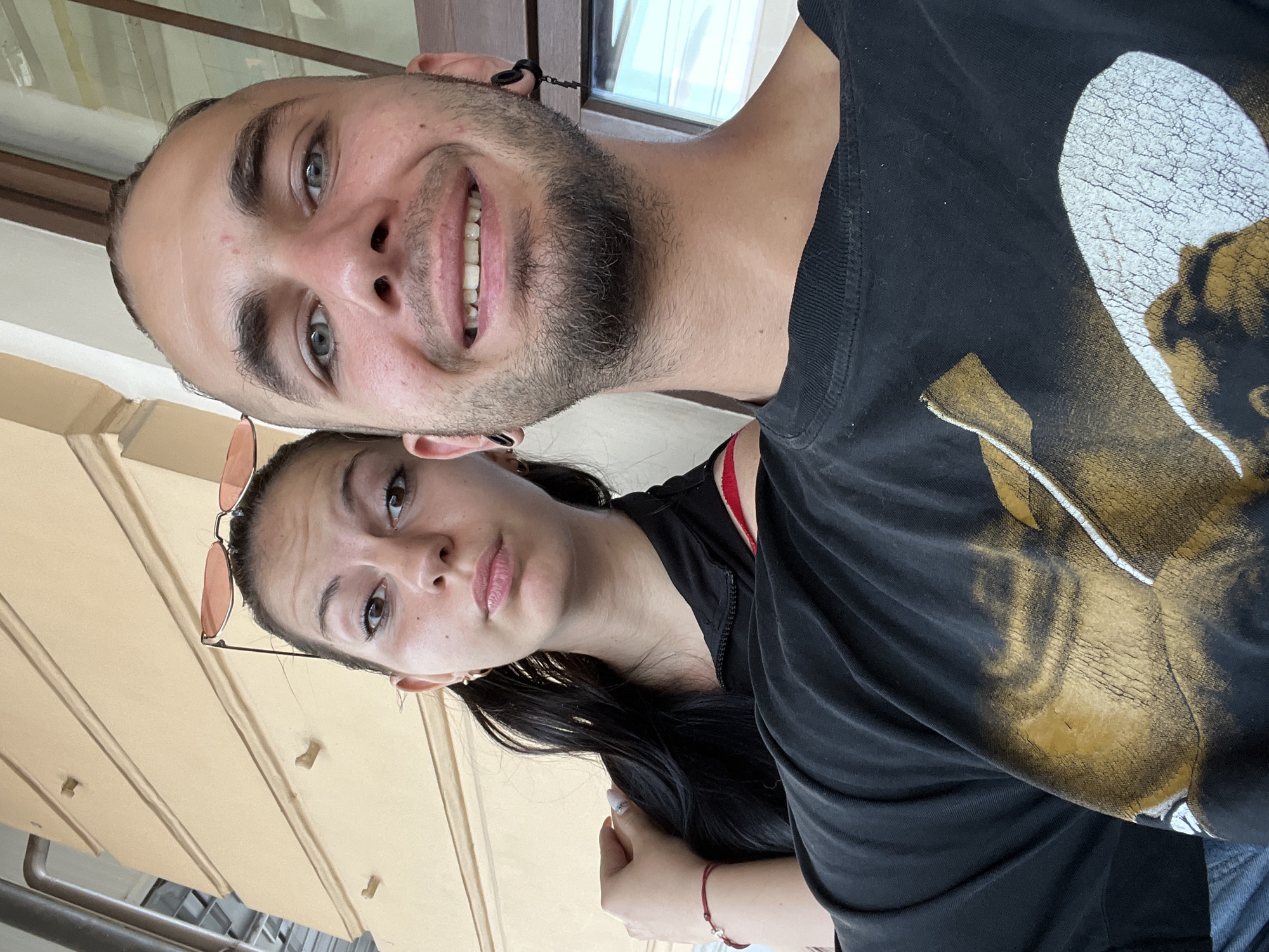 A casual selfie captures two young adults, a man and a woman, in what appears to be an outdoor or semi-outdoor setting in Plovdiv, Bulgaria. The image is taken at a Dutch angle, rotated about 90 degrees counter-clockwise.
In the foreground, a man with a short beard and light-colored eyes smiles broadly, looking directly at the camera. He wears a black t-shirt featuring a distressed graphic design in white and gold/brown, which appears to depict a stylized figure or face. Dark ear tunnels are visible in his earlobes.
Behind and slightly below him, a woman with long dark hair and brown eyes looks towards the camera with a neutral, slightly pouty expression. She has sunglasses perched on top of her head and is wearing a black top, with a glimpse of a red strap visible underneath. A red string bracelet with a small charm is on her wrist, and her nails are painted light with a design.
The background shows elements of a building, including a light brown/beige facade with horizontal paneling and a window reflecting bright light. A metal railing is partially visible at the bottom of the frame. The bright lighting indicates it is daytime. The overall mood conveyed is relaxed and informal.