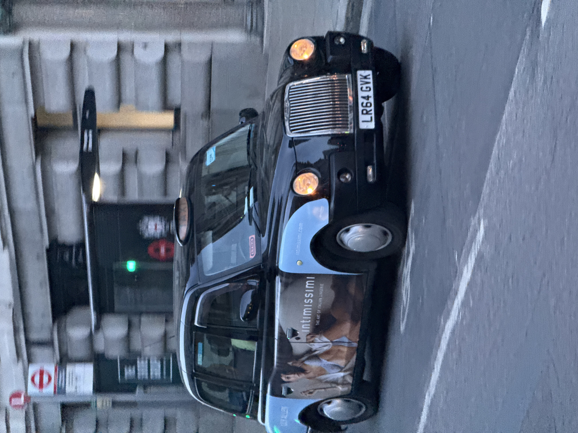 The image shows a black London taxi cab, with the license plate LR64 GVK, on a city street. The taxi has an advertisement for Intimissimi lingerie on its side. The background indicates the street is in London, based on the architecture. A pedestrian crossing sign is visible.