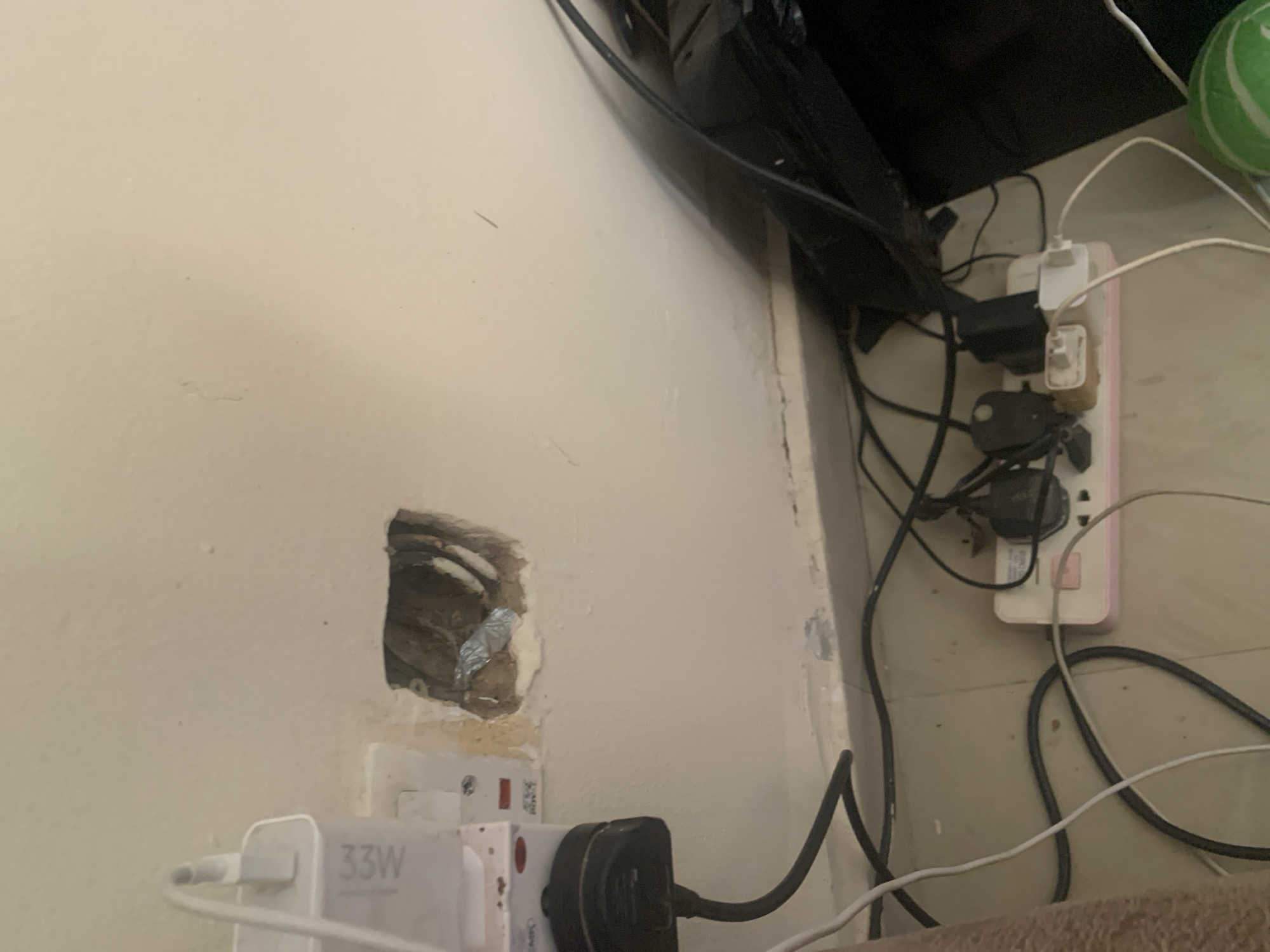 In Gura Top, Nigeria, a close-up shot reveals a section of a wall with damaged plaster, exposing electrical wiring within a hole. Adjacent to this is a white electrical outlet with a charger plugged into it, labeled "33W". Below the damaged wall section, a pink power strip is visible on the floor, connected to multiple electronic devices via various adapters and plugs. The floor is tiled and a black backpack or bag is partially visible on the right side. The scene appears to be in a residential or office setting, with multiple electronic devices and charging apparatuses.