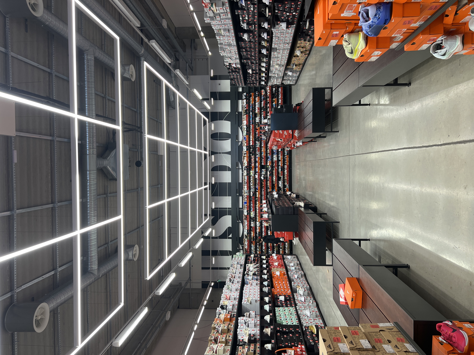 The image captures an interior view of a retail establishment, likely a shoe store, specifically a Nike outlet. Rows of shelves laden with shoe boxes extend into the distance, their colors ranging from vibrant hues to neutral tones, meticulously arranged by size and type. A broad stripe of black color runs along the centerline of the image, and the word "SPORTS" in large white sans-serif letters is prominently visible along the central axis, presumably an architectural feature of the space.
Illuminating the scene are square LED lights, casting a bright glow on the merchandise and pathways. The ceiling features industrial-style ductwork and support structures.
The location in the city of Unknown, Unknown cannot be determined based on this image.