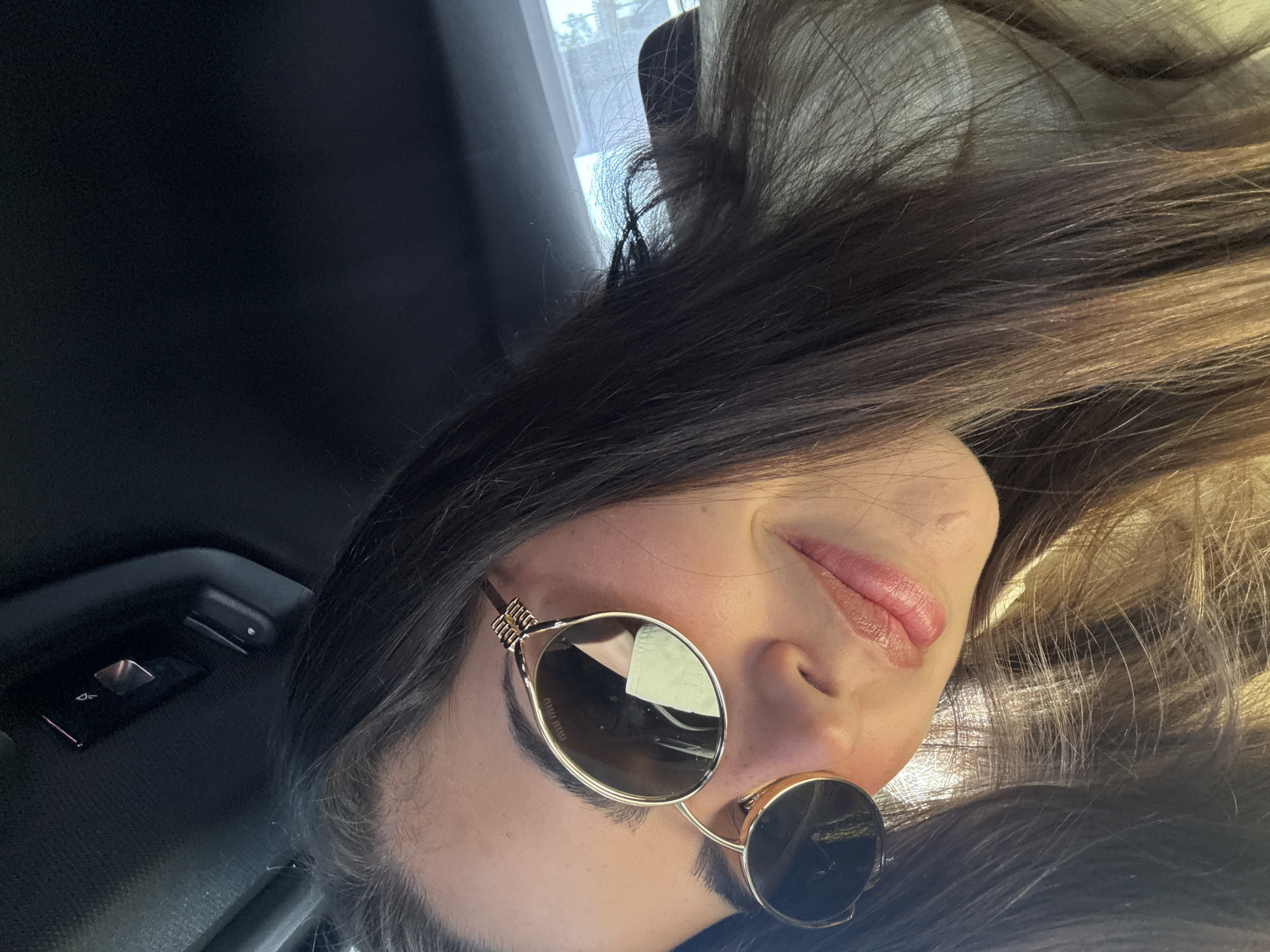 A woman is pictured indoors, likely in a car, based on the interior features visible in the top left corner. She is wearing round sunglasses with gold frames. Her hair is long, brown, and flowing around her face. The background is blurred, with a hint of a window or light source at the top. There is no definitive evidence in the image to ascertain that this photograph was taken in Mexico City, Mexico.