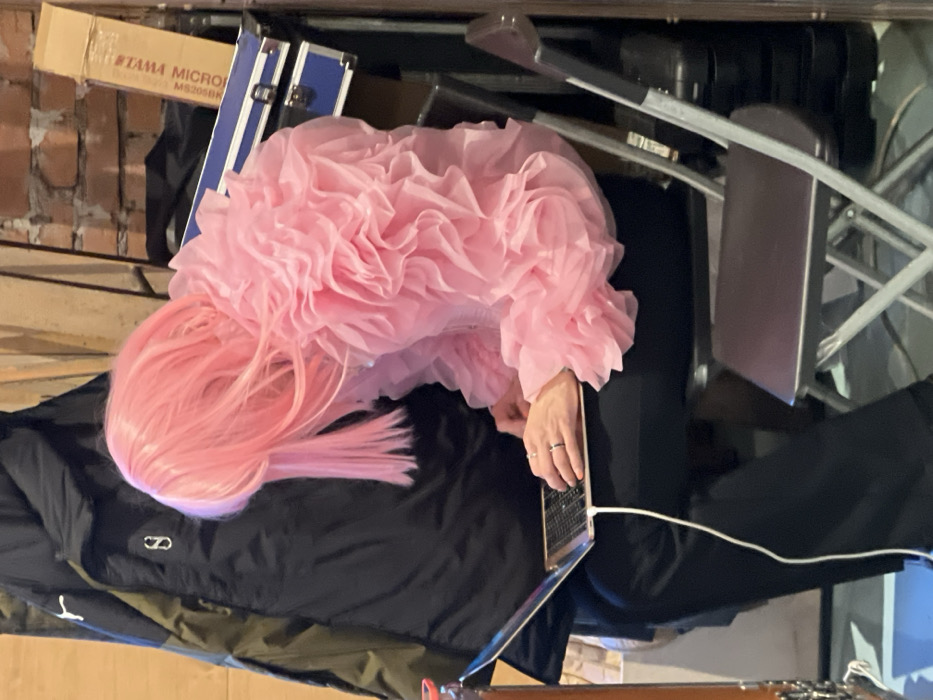 An individual, visible from the left side, is seated on a dark folding chair, facing forward with their head angled downwards. They are wearing a bright pink shoulder-length wig that obscures their face, a voluminous pink ruffled top with sheer layers, and dark trousers. Their hands are positioned over the keyboard of an open silver-colored laptop, which has a white cable connected and extending downwards. A metallic ring is worn on the ring finger of the left hand. To the immediate left of the individual, a dark winter jacket with a white "S" or "8" logo hangs against a light wooden wall. To the right and behind the individual, a reddish-brown brick wall is visible. Stacked against the brick wall are two blue hard cases, with a cardboard box positioned on top, partially displaying the text "TAMA MICRO MS205BK". The scene takes place indoors, suggesting a backstage area or studio environment. The precise location within the city of Saint Petersburg, Russia, cannot be determined from the visual evidence provided.