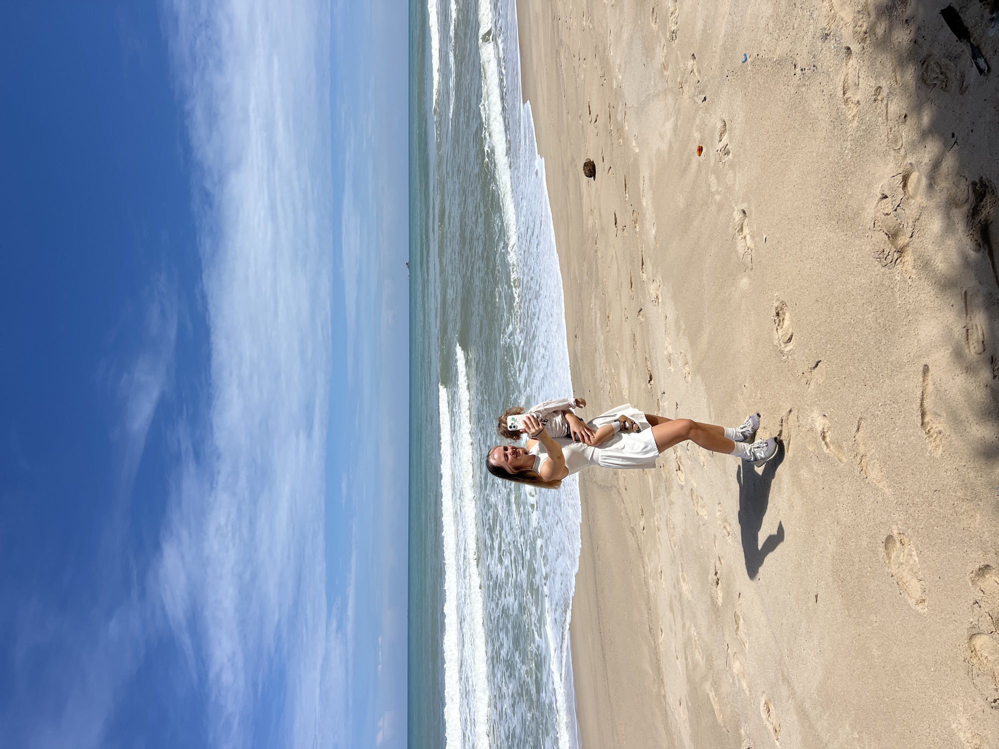 A woman in a white dress and sneakers stands on a sandy beach, holding a child in her arms. She is taking a photo with her phone. The ocean with waves is in the background, under a partly cloudy blue sky. There are footprints in the sand, and a coconut is visible near the water's edge. A small boat is in the distance on the ocean.