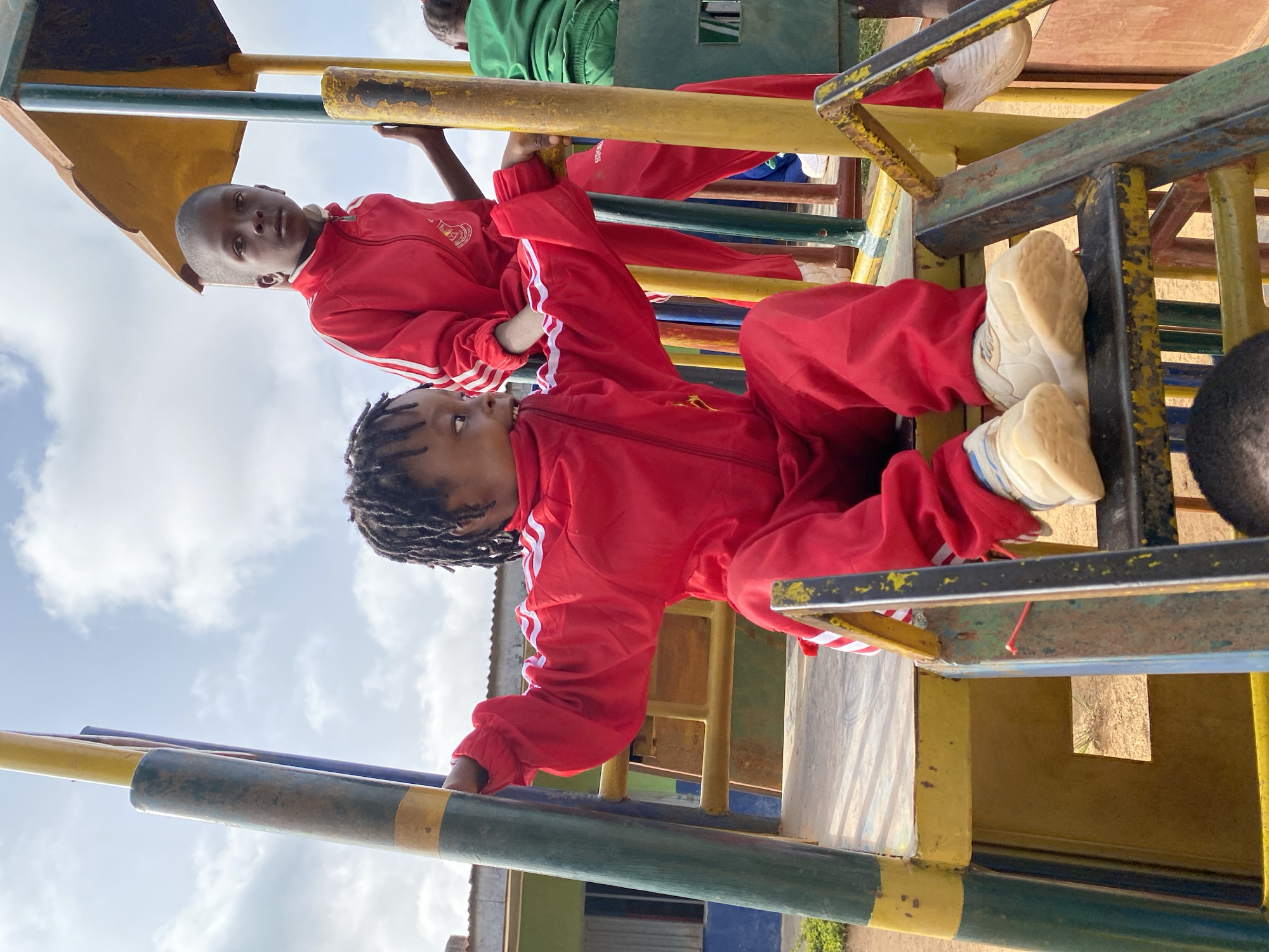 Two children are on a playground climbing structure in Gura Top, Nigeria. Both children are wearing red tracksuits with white stripes on the sleeves and white sneakers. One child is closer to the camera, hanging from a bar, while the other is above and slightly behind them. The playground equipment is constructed from painted metal bars in yellow, green, and blue. The sky with some clouds is visible in the background. The location appears to be outdoors, near a building with a light green and blue exterior.