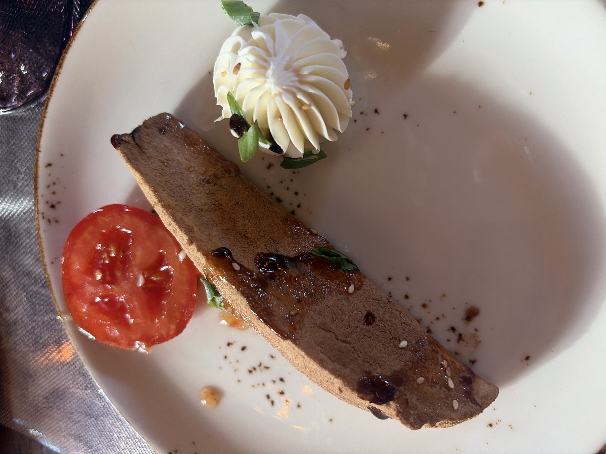An overhead, close-up shot features a meticulously plated appetizer on a white, round dish with a textured, brownish rim. Dominating the center is a long, rectangular piece of what appears to be pâté or a savory terrine, characterized by its smooth, brown texture. It is artfully garnished with a dark, glistening glaze, small green herb leaves, and scattered white sesame seeds.
To the upper right, a decorative swirl of white, creamy substance, resembling piped butter or a soft cheese spread, stands out. This swirl is adorned with small green leaves and tiny, light-colored seeds, possibly pine nuts or sesame seeds. A fresh, bright red tomato slice, revealing its inner pulp and seeds, is positioned to the lower left of the pâté.
The white surface of the plate shows sparse dark specks, possibly ground spices or residue from the glaze. The lighting creates bright highlights on the food items, particularly the tomato and the creamy swirl, suggesting an indoor setting. Part of a dark, textured object, likely a glass or ceramic, is visible on the far left edge, along with a grey, textured fabric, possibly a tablecloth or napkin, beneath the plate. No individuals or text are visible in the image.