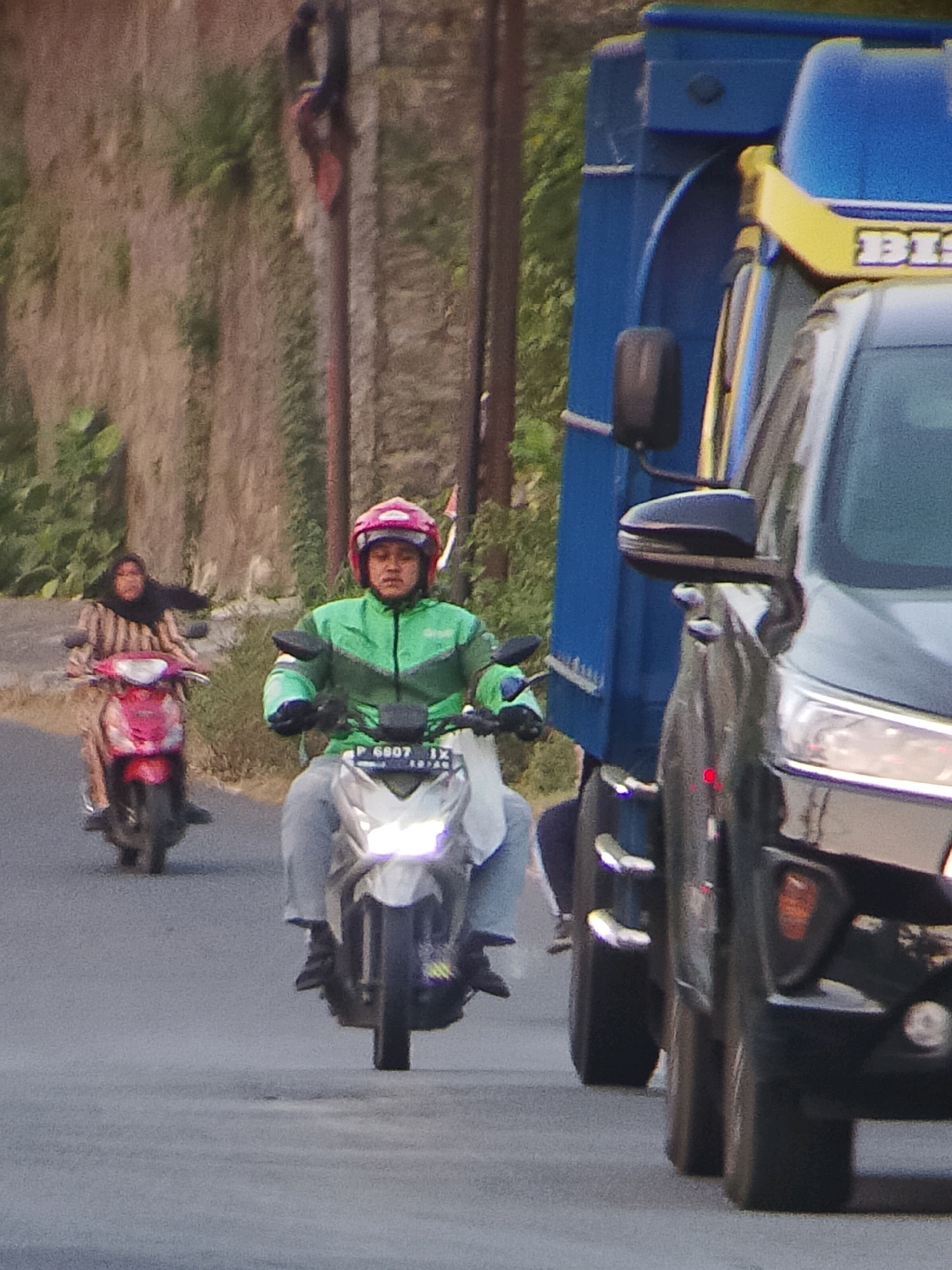 The image shows a street scene in Jember, Indonesia, with several vehicles and people. In the foreground, a person wearing a green jacket and a pink helmet is riding a white scooter, their face is visible and they appear to be looking forward. The scooter has a license plate that reads "P 6807 JX". To the left of this scooter, another scooter with a rider wearing a brown and white striped hijab is visible, the scooter is pink. In the background, a large blue truck is partially visible on the right side of the image. The truck appears to be a cargo truck. The road is asphalt and there is a dry, vegetated embankment on the left side of the road. The lighting suggests it is daytime, possibly late afternoon.