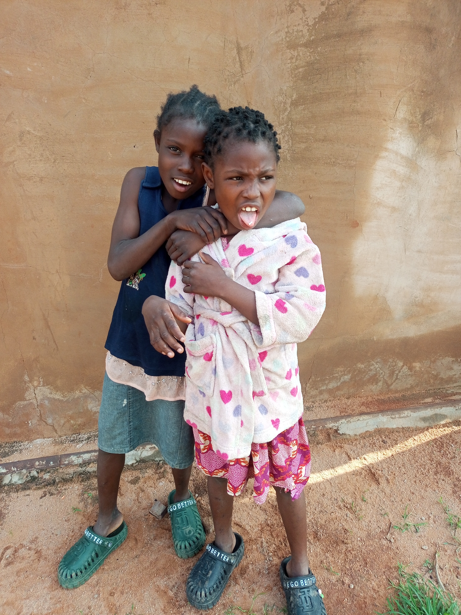 Two young girls stand in front of a rough, tan-colored wall. The girl on the left wears a sleeveless dark blue shirt and a light blue denim skirt, her arm around the other girl's shoulders. The girl on the right wears a pink bathrobe patterned with hearts and a colorful skirt. She has her tongue sticking out and is looking to her right. Both girls have dark skin and braided hair, and they are both wearing clogs. The ground is covered in reddish-brown dirt, with patches of dry grass and a metal pipe visible. The lighting suggests it is daytime. This scene appears to be in an outdoor urban or rural area, possibly in Tempure, Nigeria, given the environmental cues.