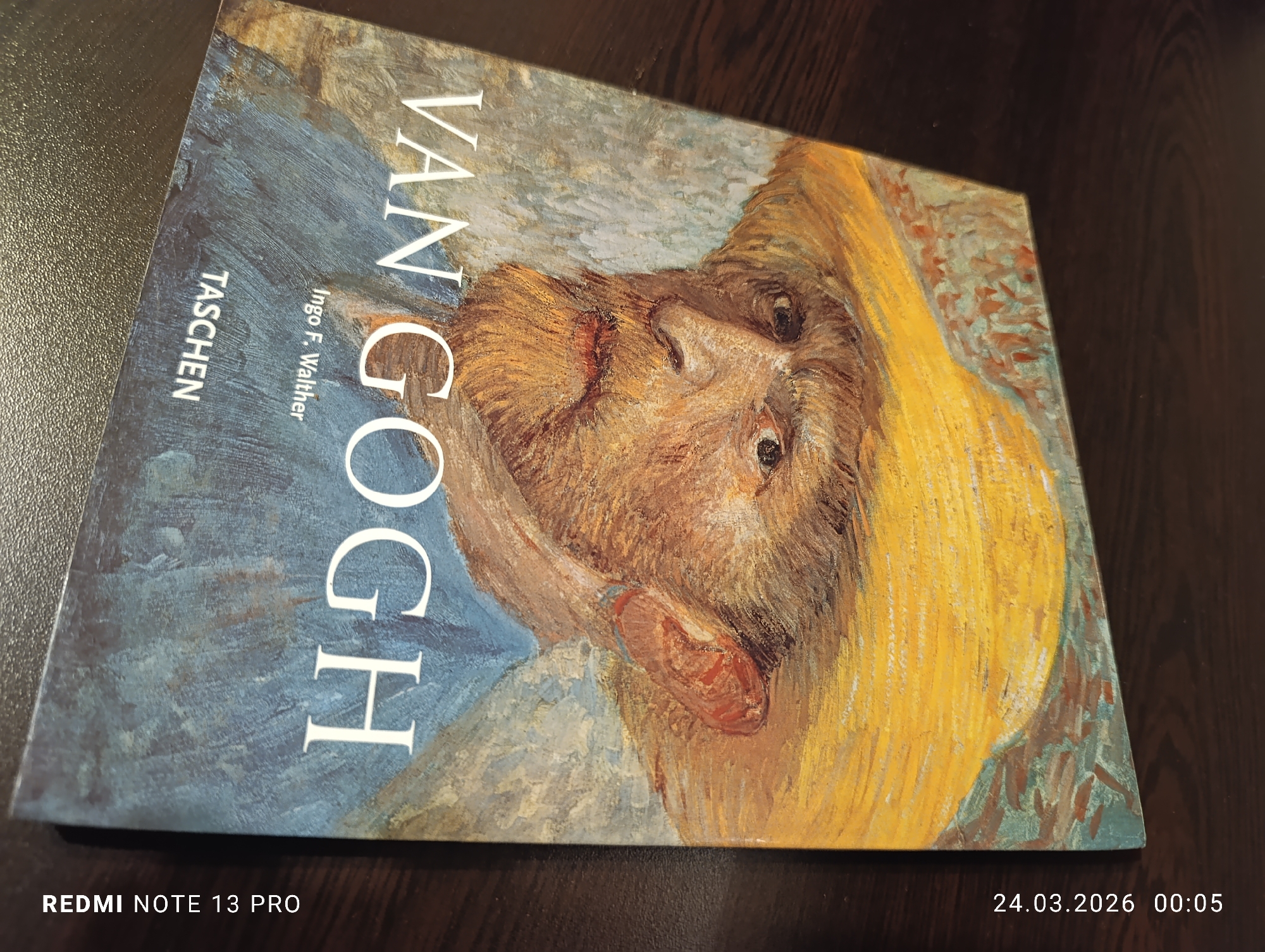 A large format art book titled "VAN GOGH" by Ingo F. Walther, published by TASCHEN, rests on a dark, patterned surface in an indoor setting in Varna, Bulgaria. The book's cover displays a reproduction of a self-portrait painting of Vincent van Gogh, depicting a man with an orange-brown beard, wearing a yellow hat or scarf, and featuring a prominent bandage over his right ear. The painting's background consists of a textured blend of blue, grey, and brown tones. The title "VAN GOGH" is rendered in large white capital letters, vertically oriented along the left side of the cover. The book is positioned diagonally in the frame, with its spine running along the top-left edge. The image metadata indicates it was captured by a REDMI NOTE 13 PRO device on March 24, 2026, at 00:05.